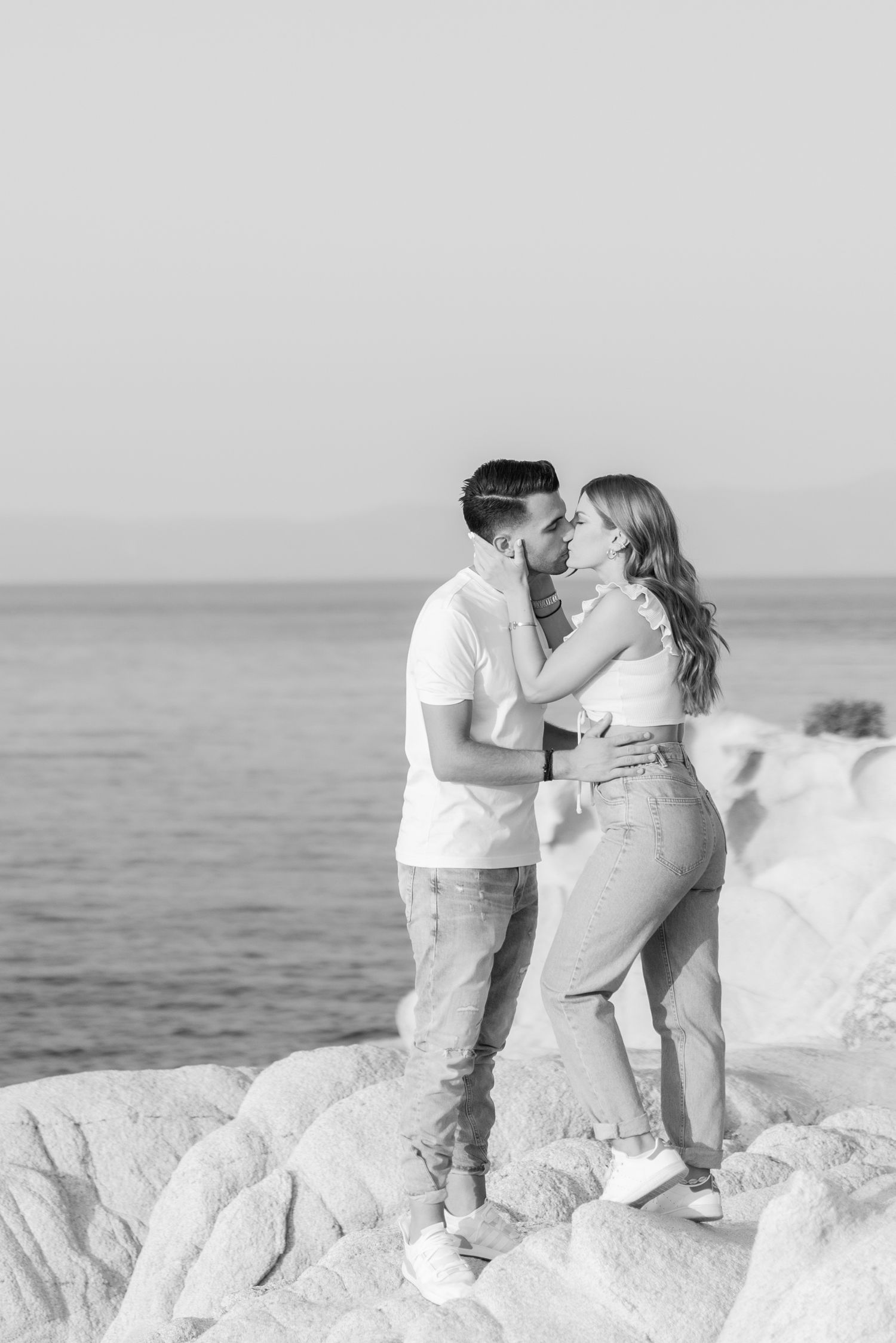 A couple shares romantic kisses on white coastal rocks overlooking the ocean.