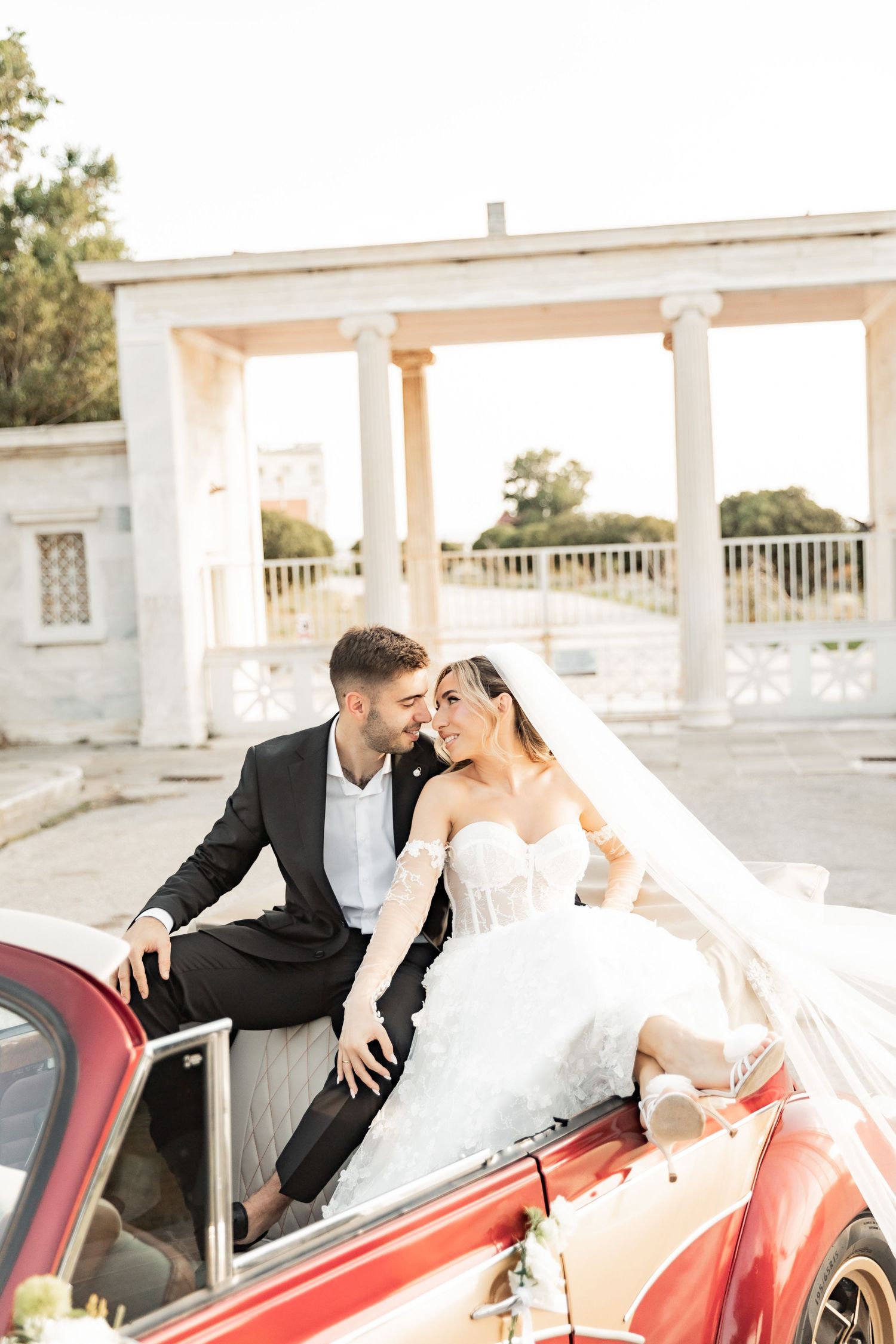 Bride and groom sitting on a vintage red car, sharing a romantic moment in front of a white classical colonnade.