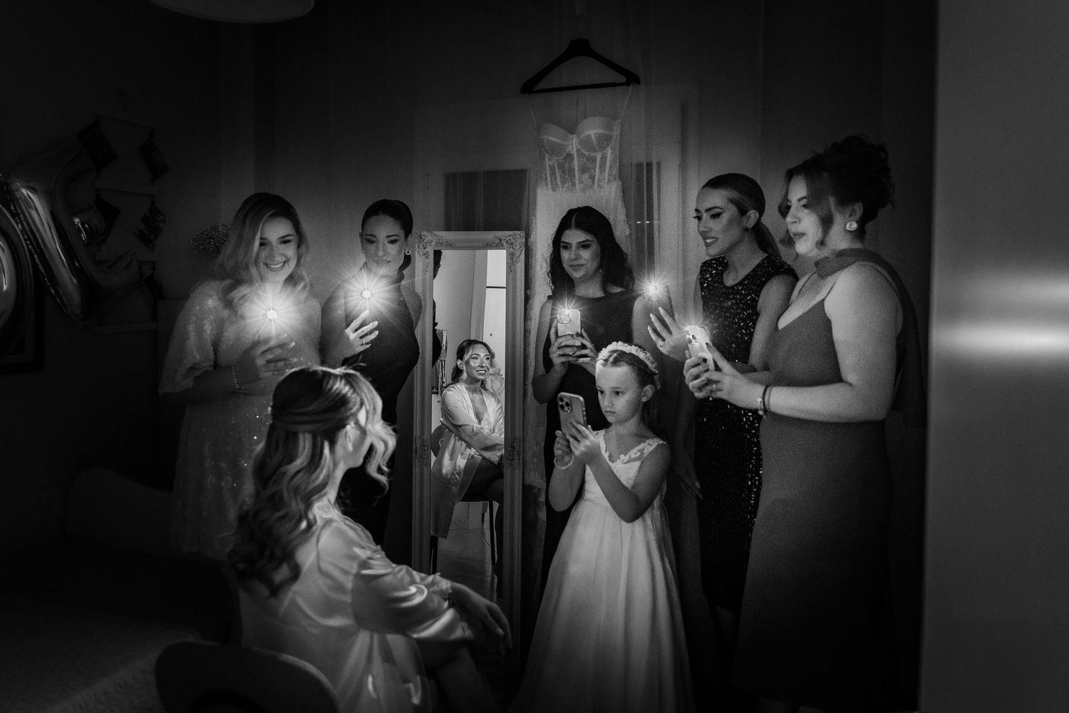 Black and white photo of bridesmaids holding phone lights around a seated bride, with a wedding dress visible in a mirror.