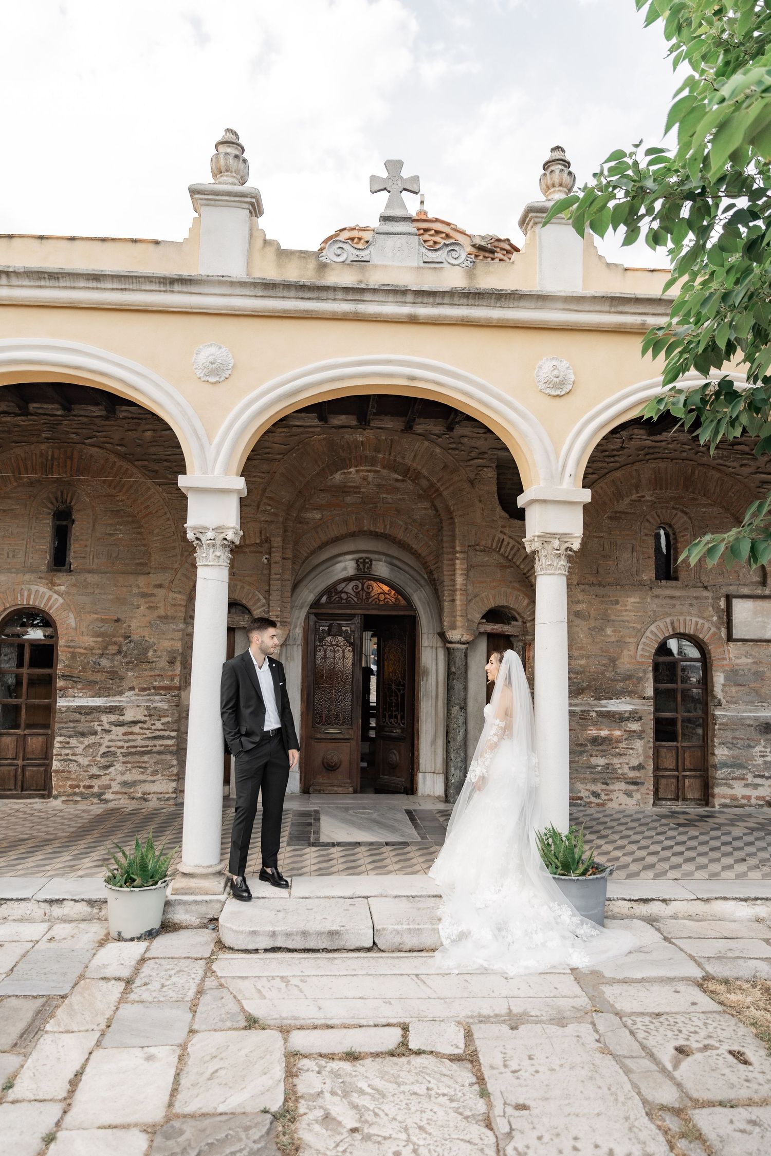 Bride and groom stand before a historic stone church with arched columns and a cross on their wedding day.