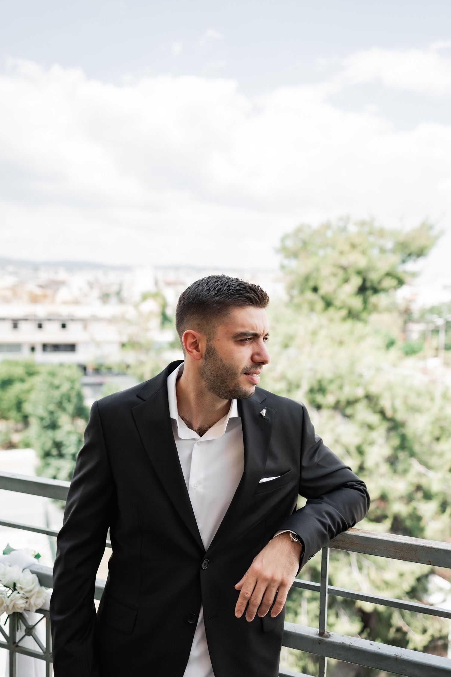A stylish man in a black suit leans on a balcony railing with a green cityscape and cloudy sky behind him.