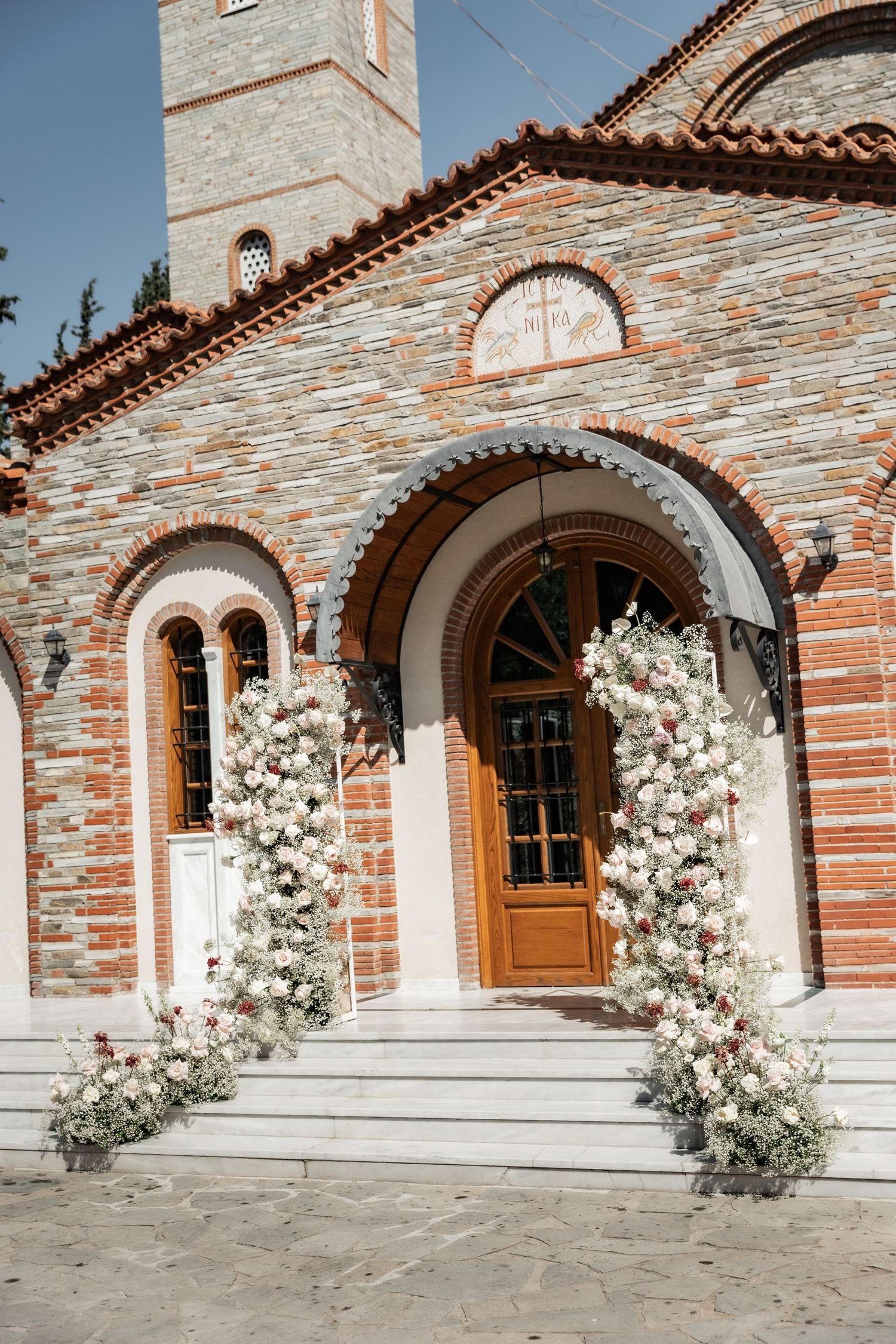Elegant brick church entrance adorned with white floral arrangements flanking wooden doors for a wedding ceremony.