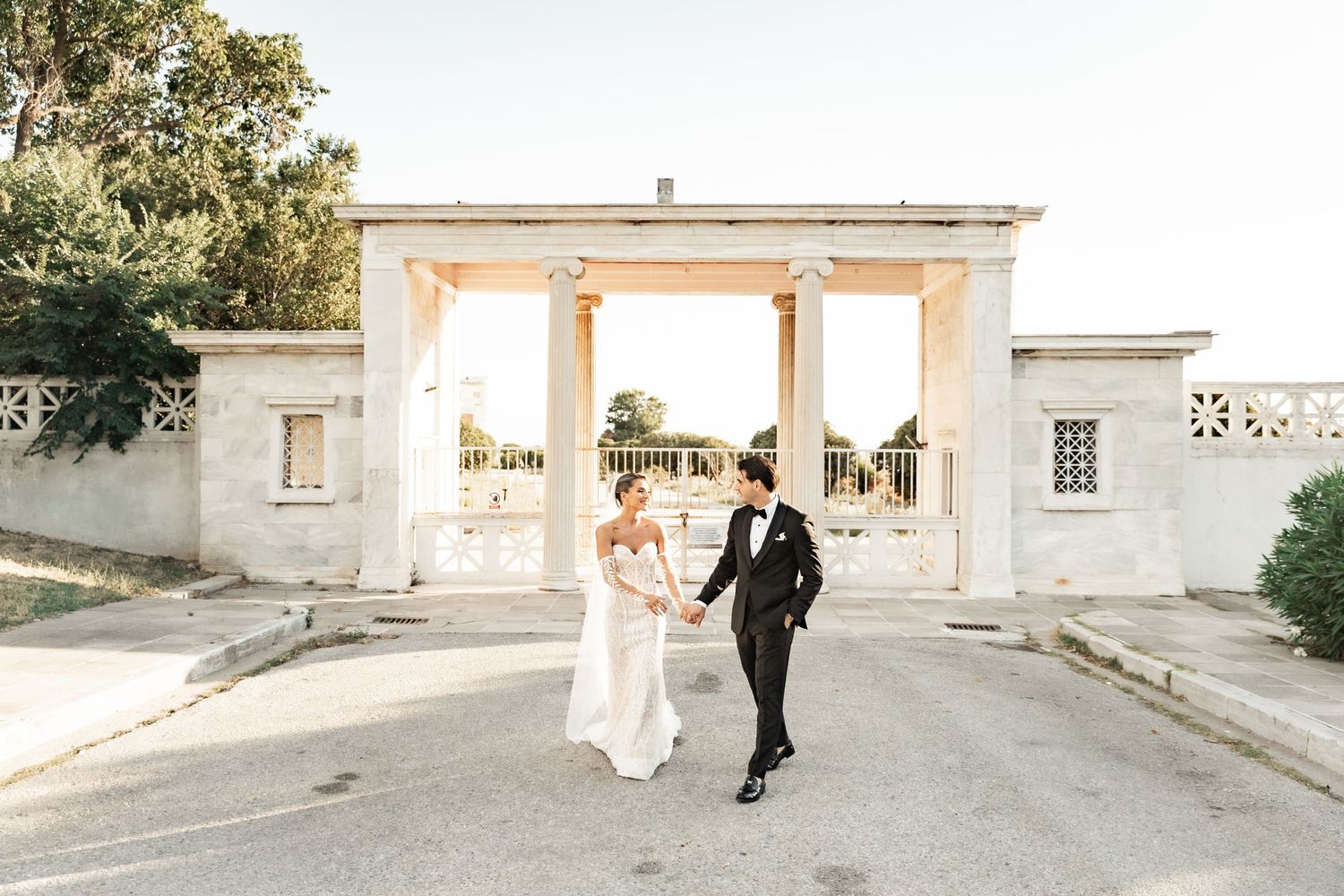 A bride and groom hold hands walking toward a grand white columned gate at a sunlit outdoor wedding venue.