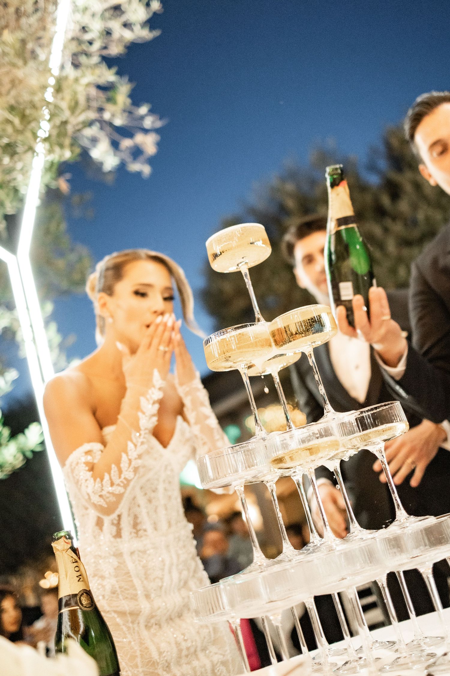 Bride and groom pouring champagne tower at elegant wedding reception under night sky.