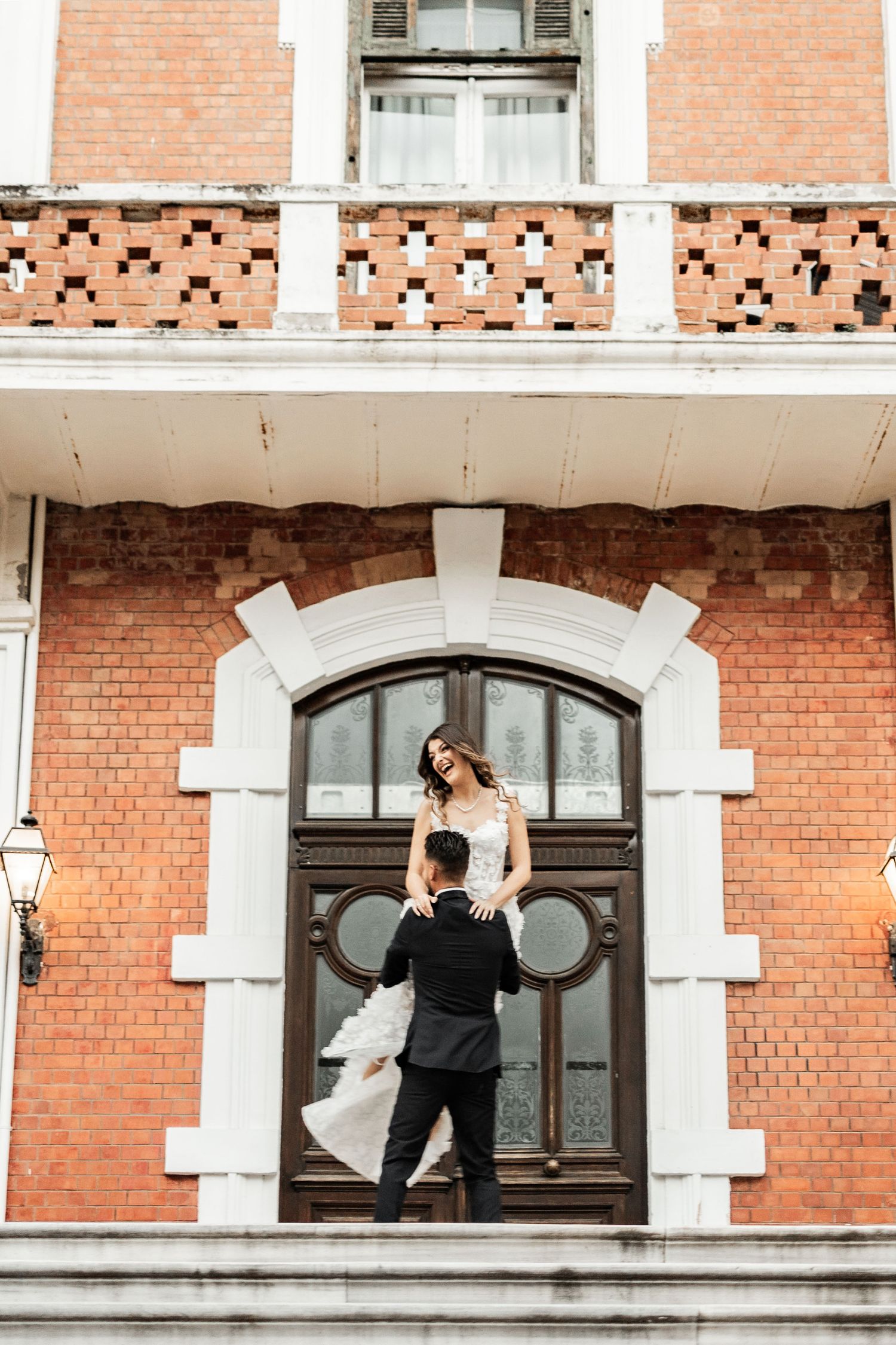 A playful wedding photo of a couple posing by an arched doorway of a brick building.