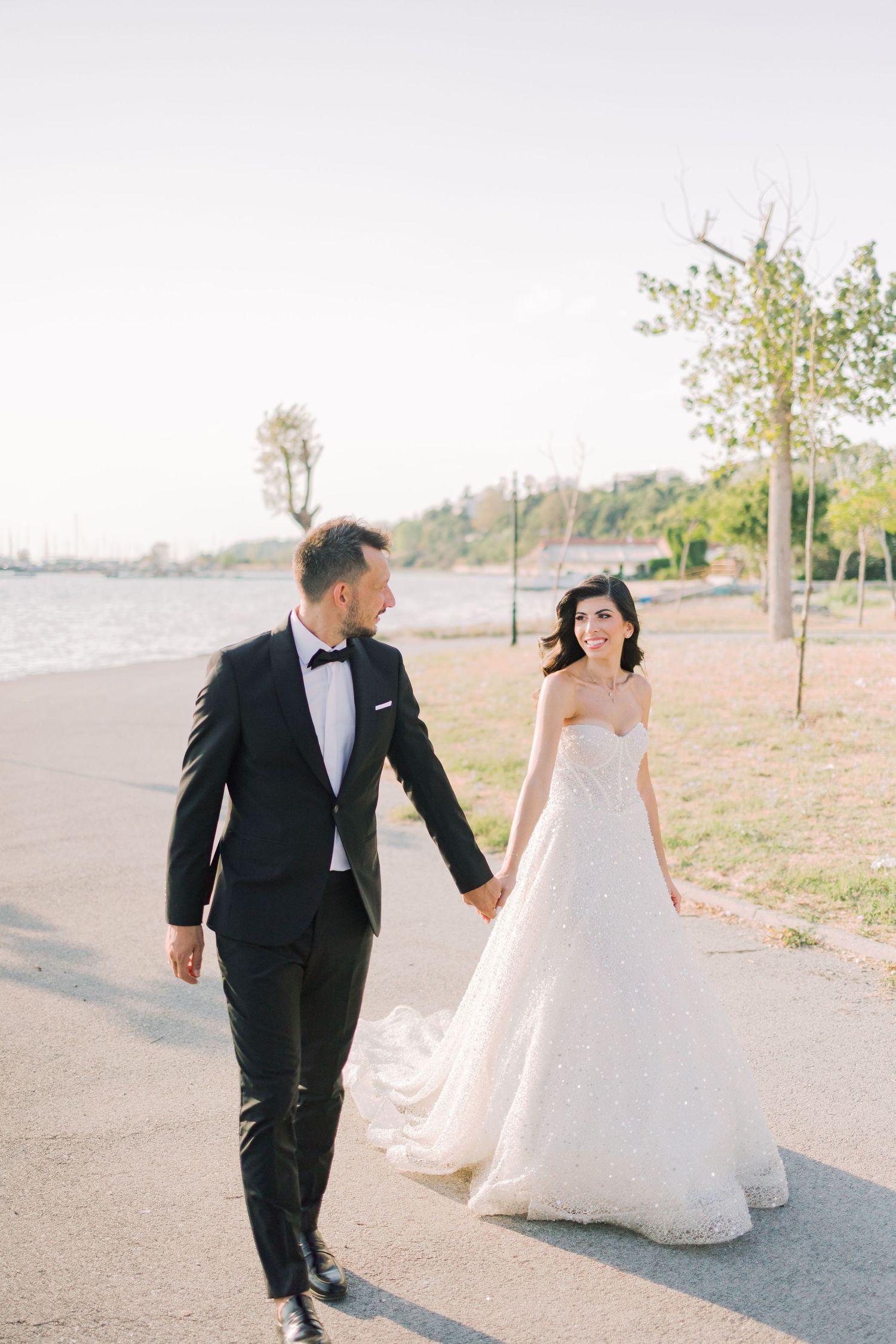 A couple walks hand in hand along a waterfront path, with the bride in a flowing white gown and groom in classic tuxedo.