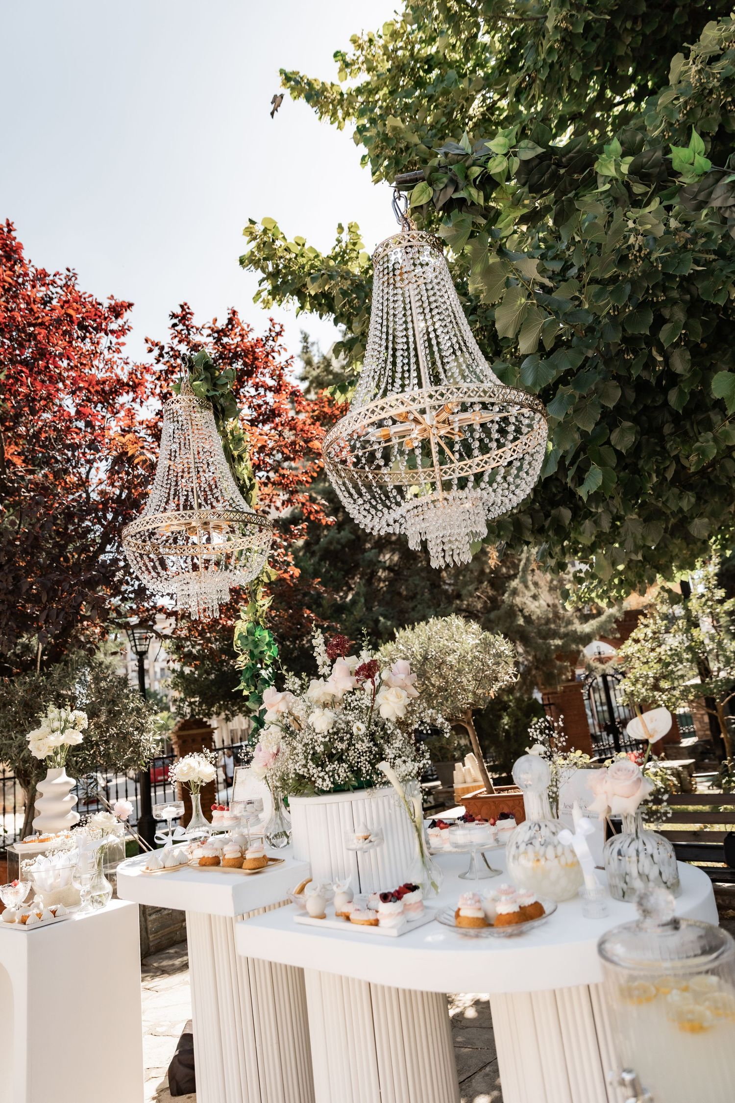 Elegant outdoor wedding dessert table with crystal chandeliers, white florals, and sweet treats under lush green and red trees.