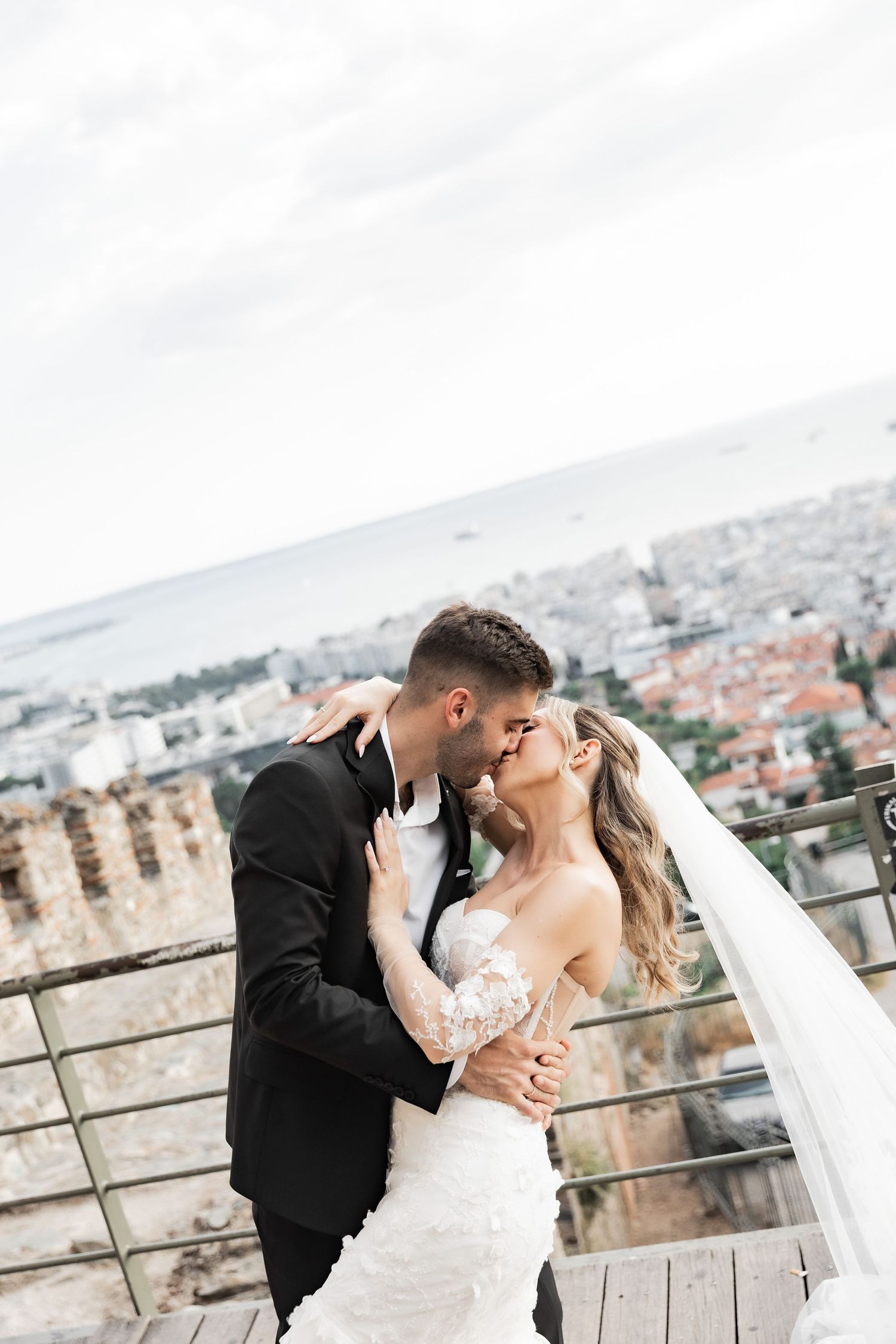 Bride and groom share a romantic kiss on a terrace overlooking a coastal city skyline on their wedding day.