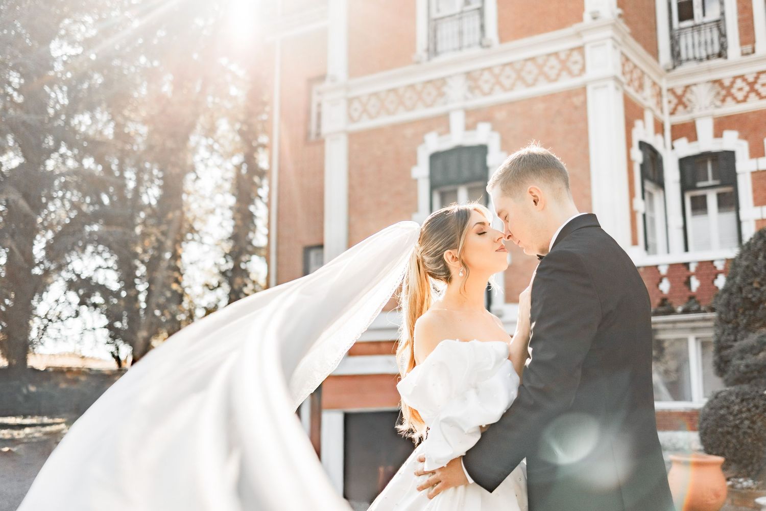 Sunlit wedding portrait with bride's veil flowing in the breeze against historic architecture.