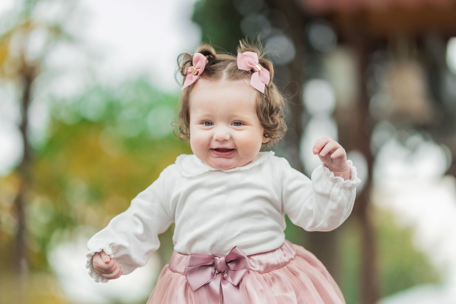 A baby in a white and pink dress smiles and plays outdoors in a garden setting.