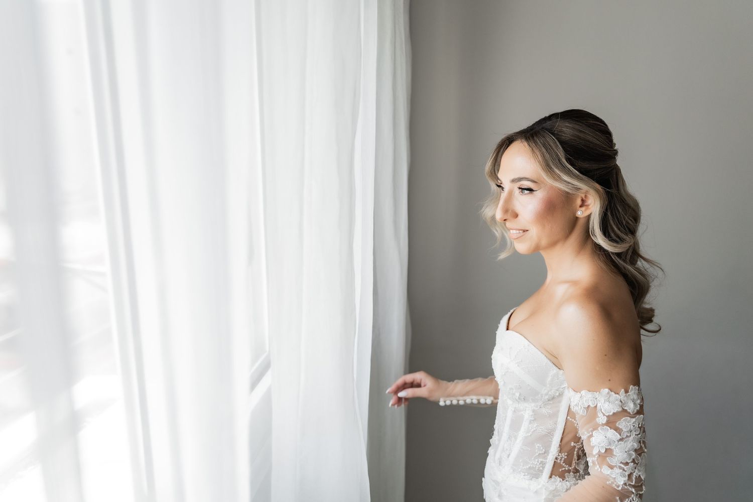Bride in lace off-shoulder wedding dress gazes out window, soft natural light, elegant half-up hairstyle.