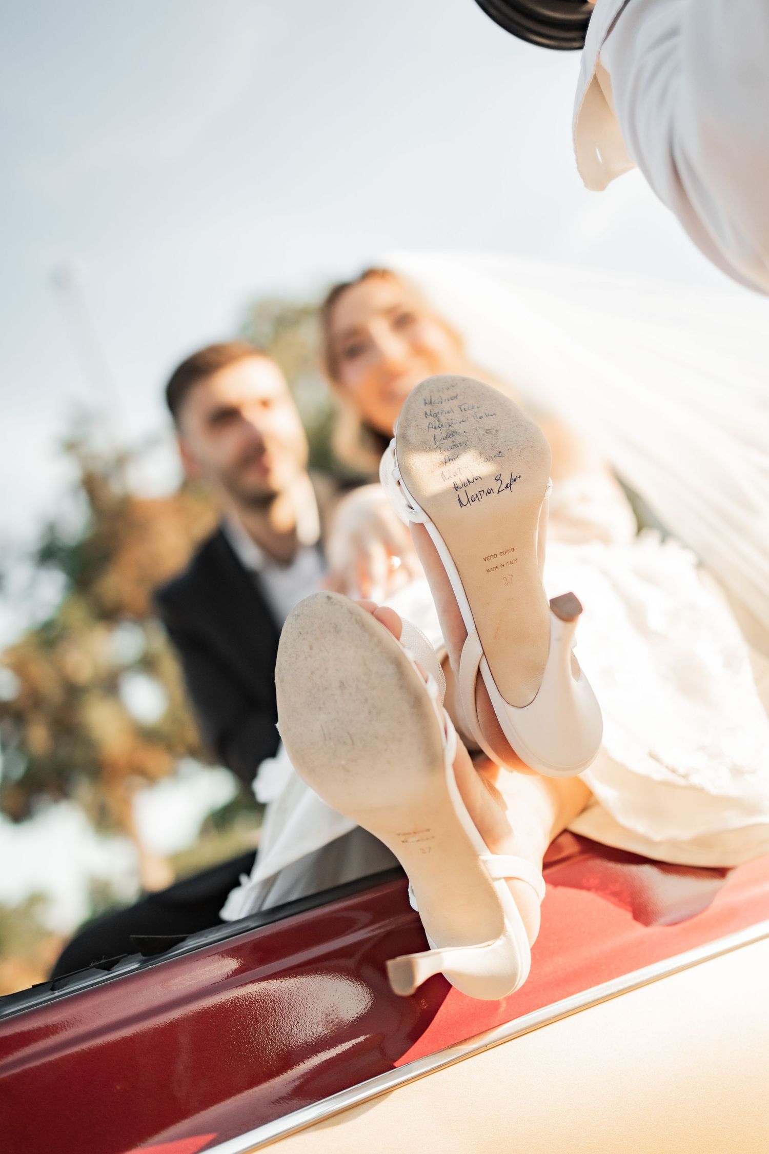 Bride and groom pose near a red car, with close-up of bridal heels showing handwritten message on soles.