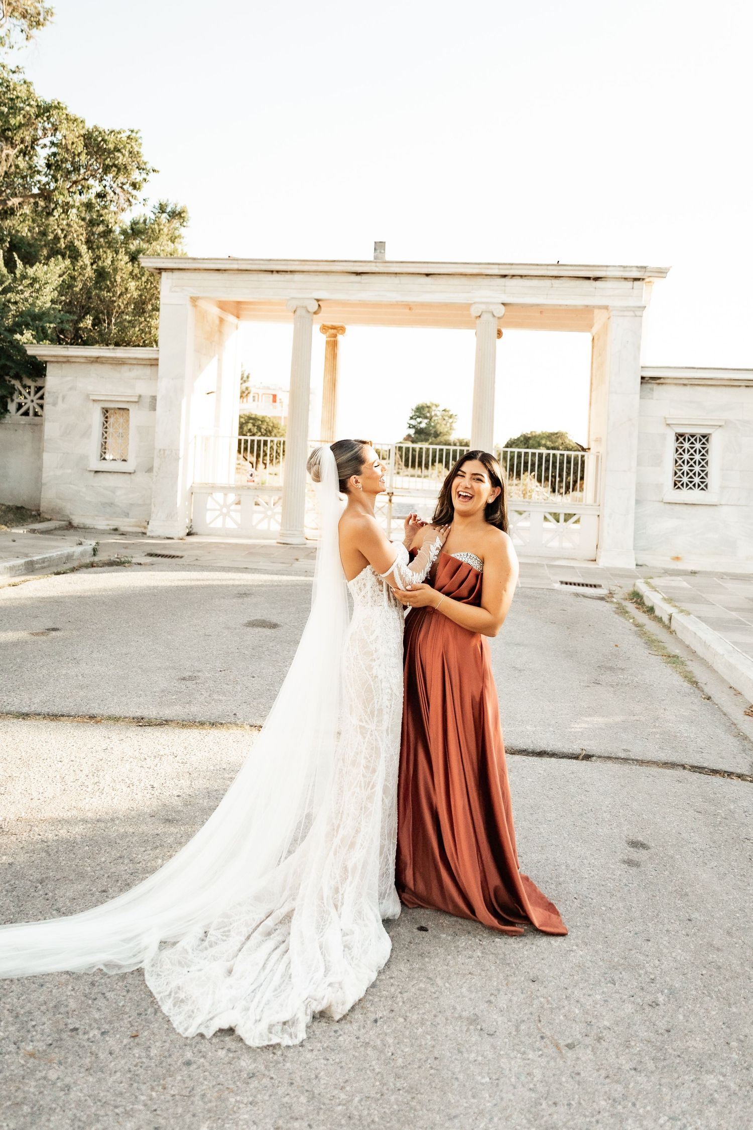 Bride in lace gown and bridesmaid in rust satin dress laughing together outside elegant white columned venue.