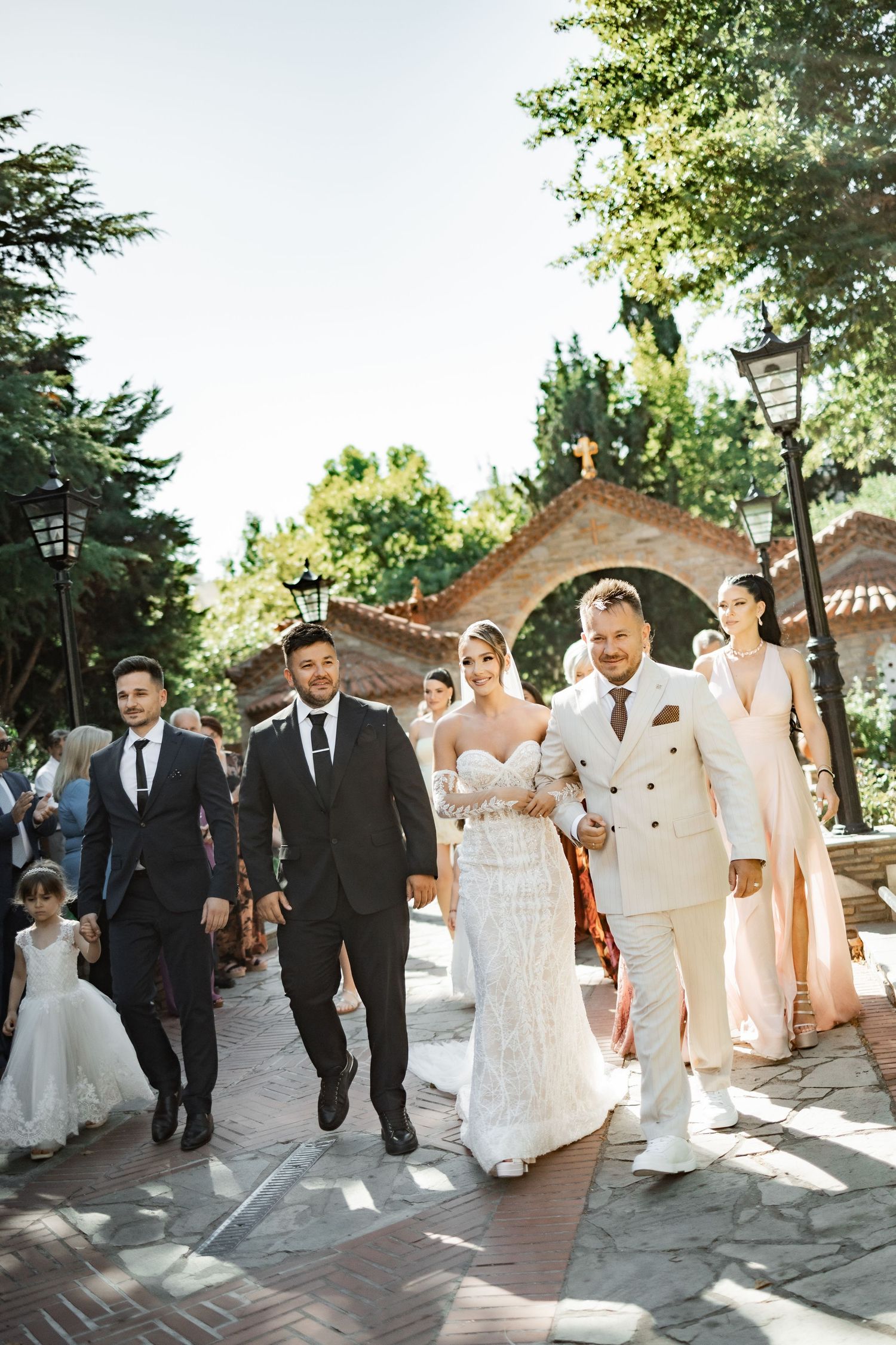 Wedding party walking outdoors near a church, bride in white gown with groom in cream suit, groomsmen in black tuxedos.