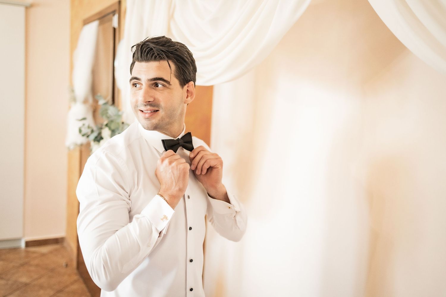 Smiling groom in white shirt adjusting black bow tie while getting ready for wedding ceremony.
