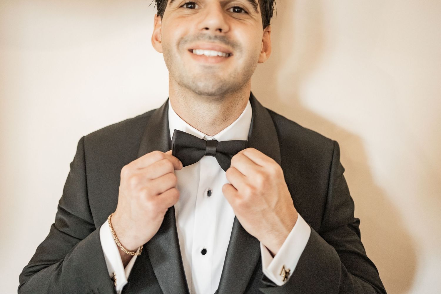 Smiling groom in black tuxedo adjusting bow tie, elegant formal wedding attire with cufflinks.