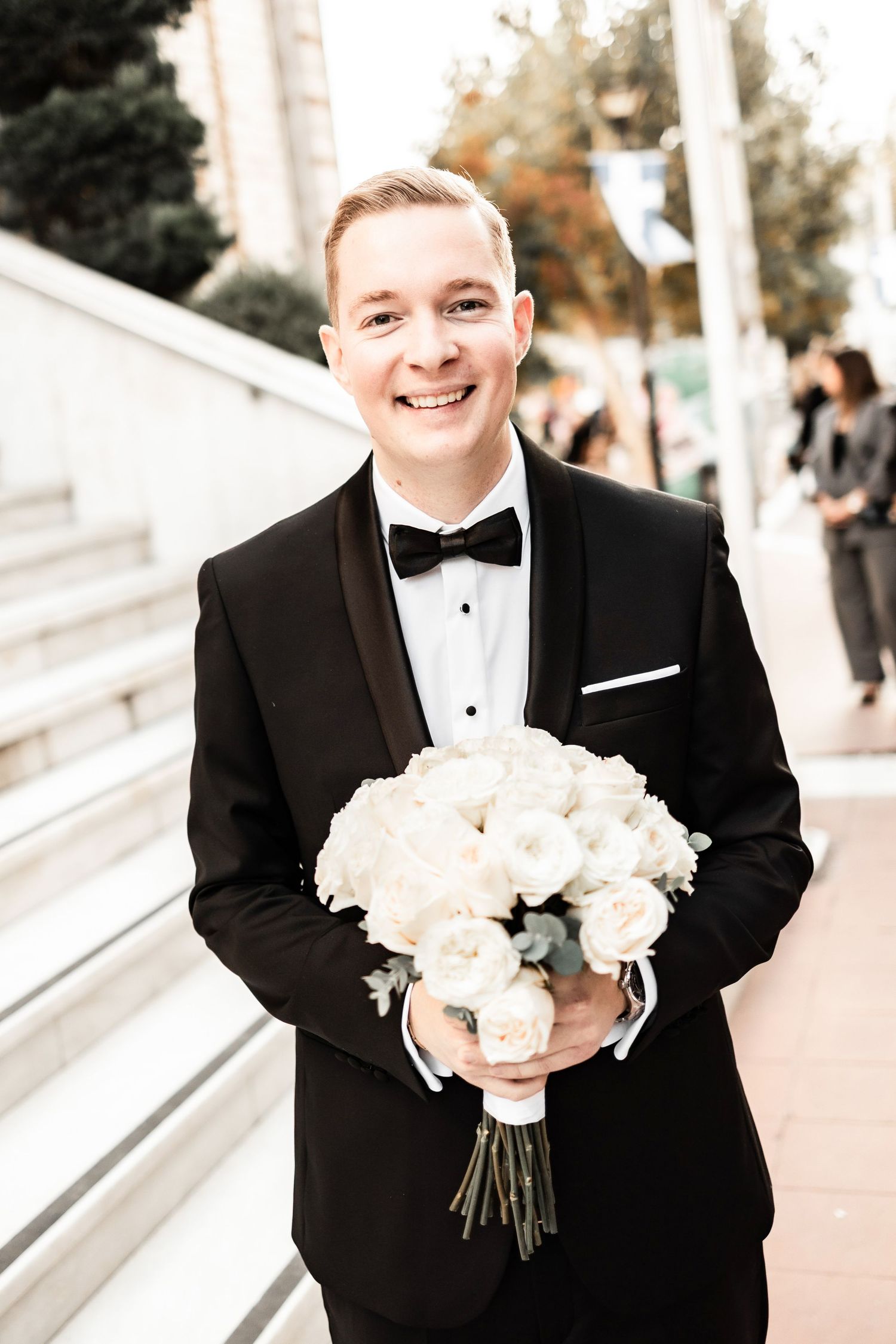 A classic wedding bouquet of white flowers held by someone wearing a black tuxedo and bow tie.