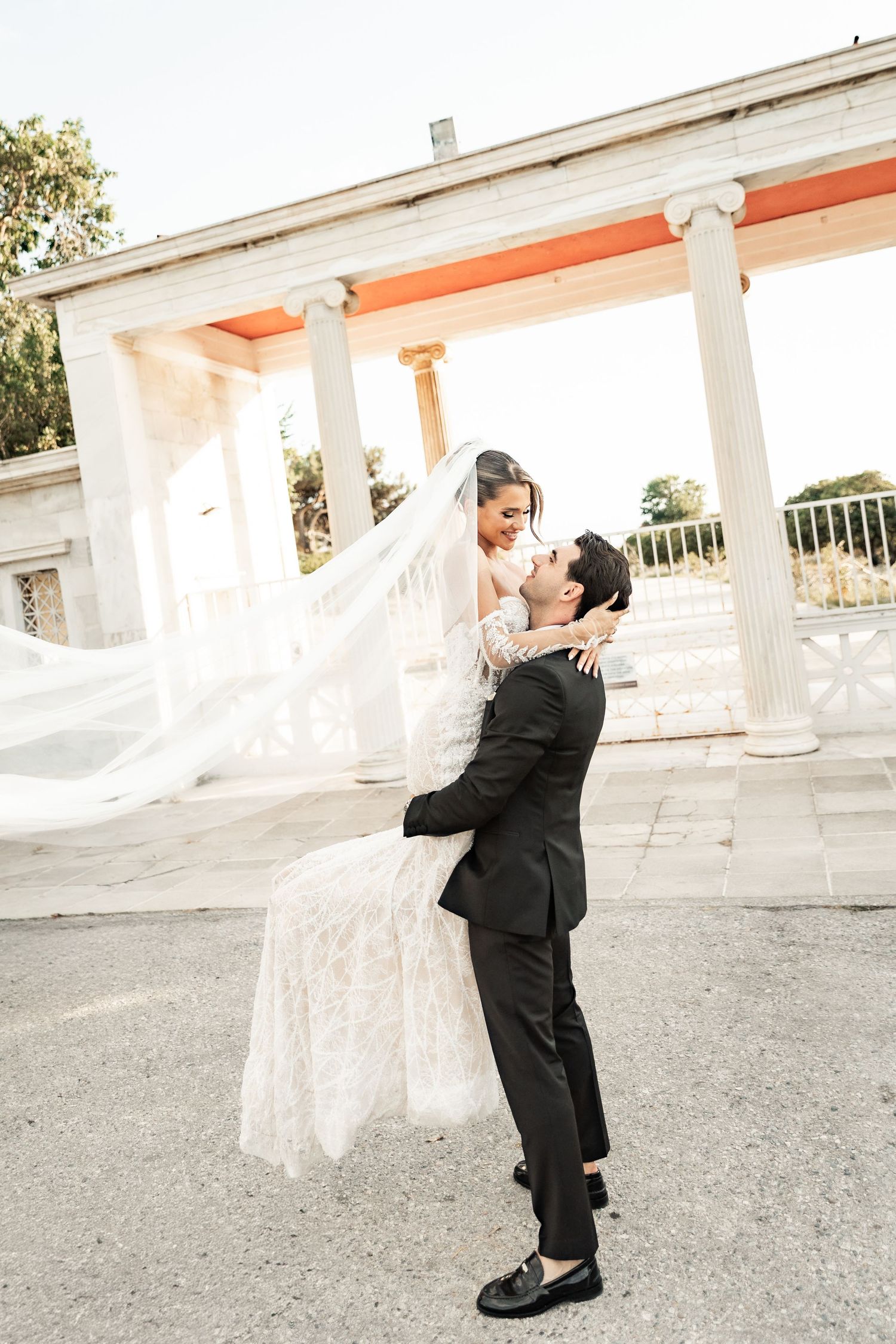 Groom lifting bride in white lace gown and veil outside elegant columned venue on their wedding day.