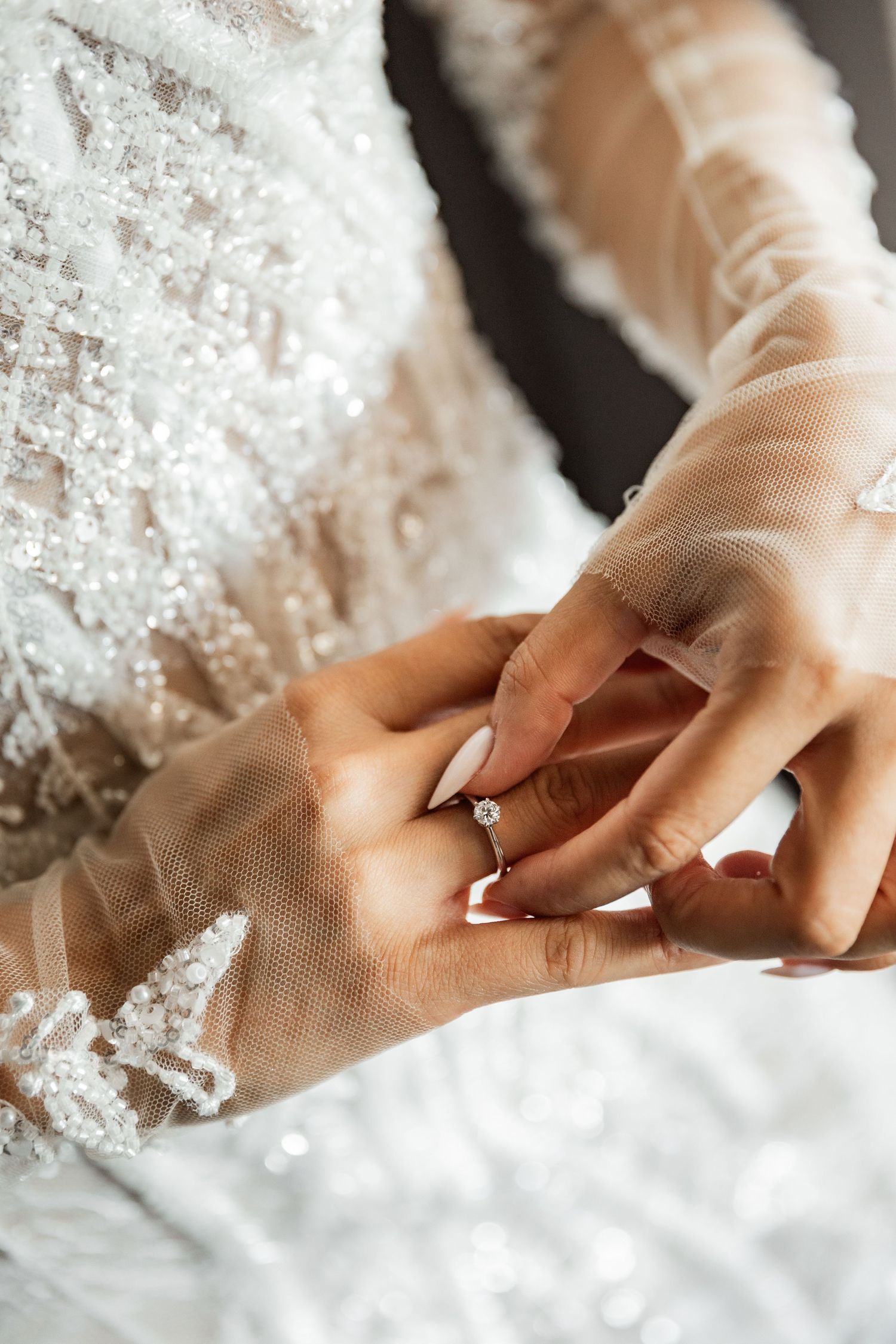 Bride in beaded white gown with sheer gloves placing a diamond solitaire engagement ring on her finger.