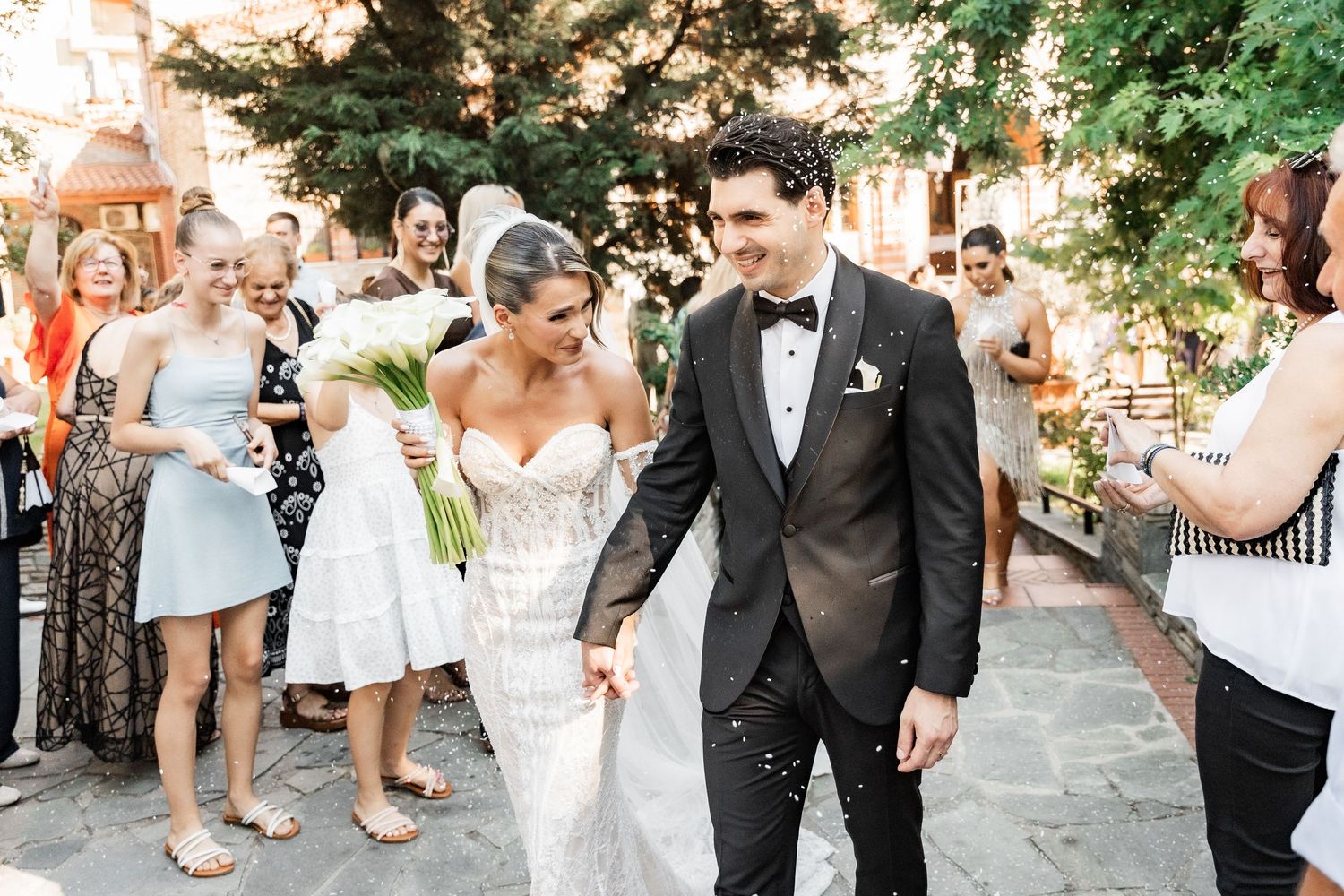 Bride and groom walk through cheering wedding guests outdoors, bride holding white bouquet in elegant lace gown.