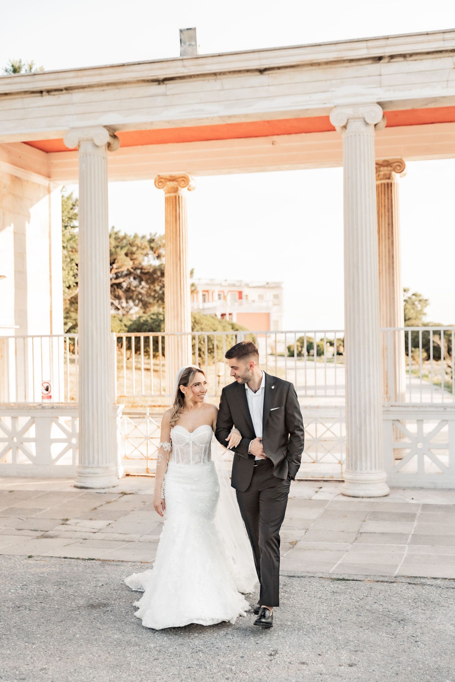 Bride and groom walking together in front of white Greek columns at an outdoor wedding venue.