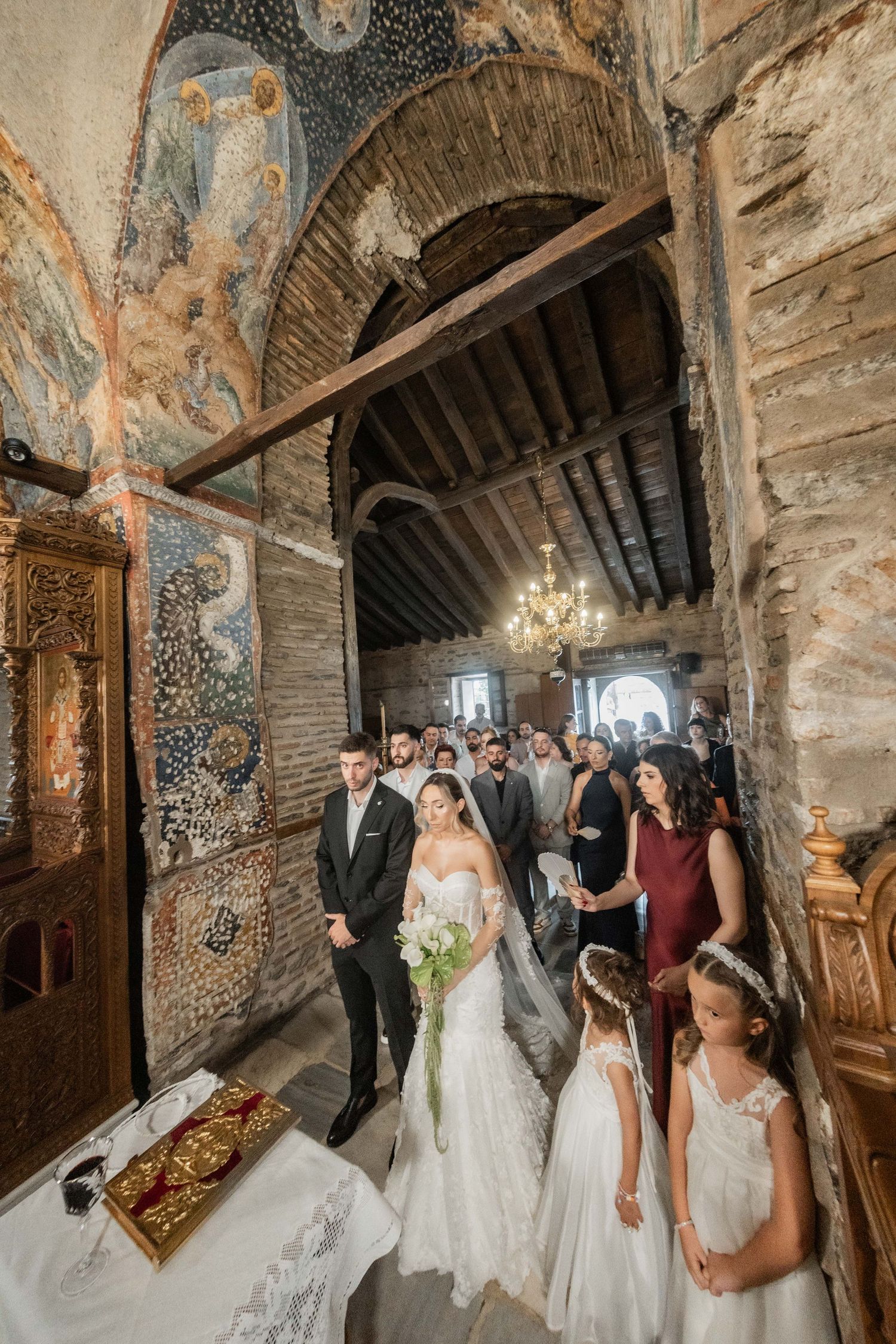 Bride and groom stand at altar in ornate Byzantine church with frescoed walls, surrounded by wedding guests and flower girls.