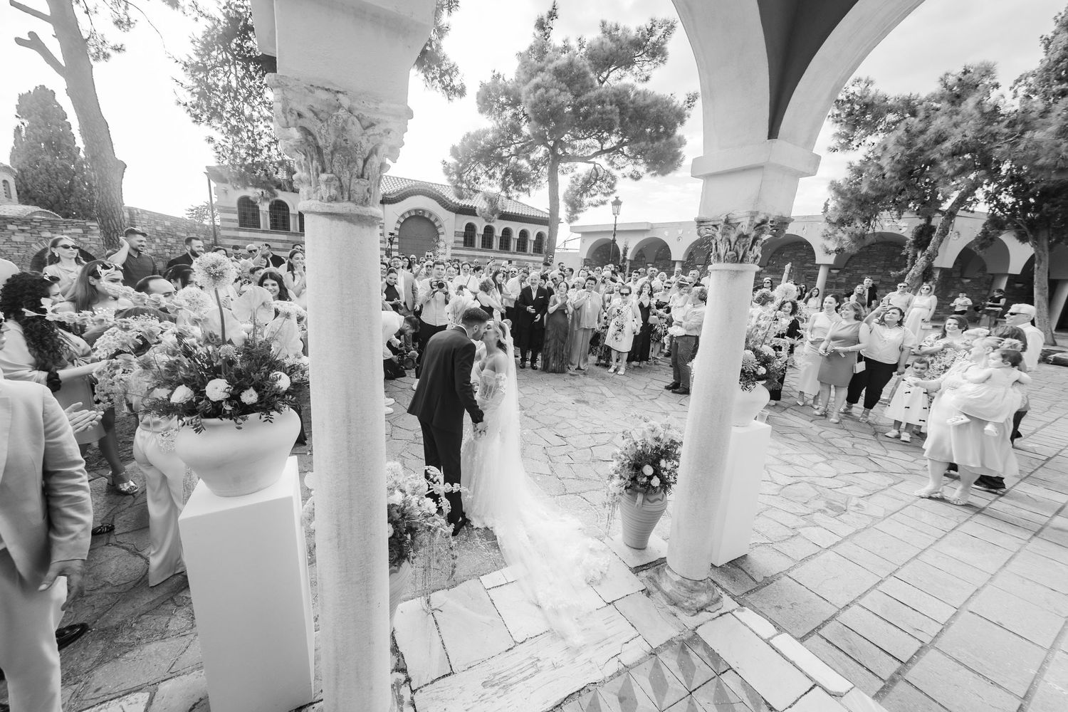 Black and white outdoor wedding ceremony with guests seated, couple at altar, floral arrangements, and elegant columned venue.