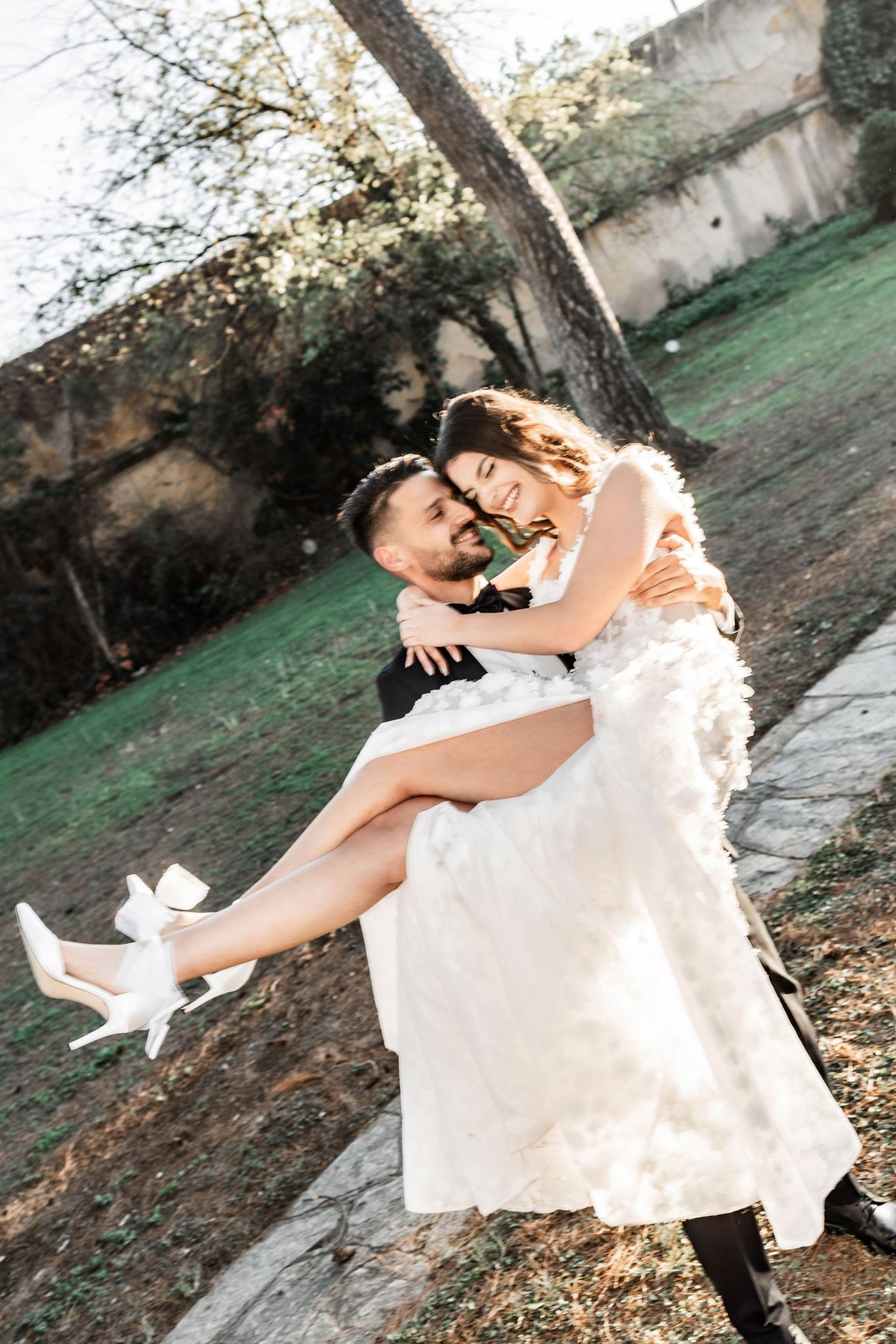 A romantic wedding photo of a couple embracing outdoors, with the bride in a white dress and heels.