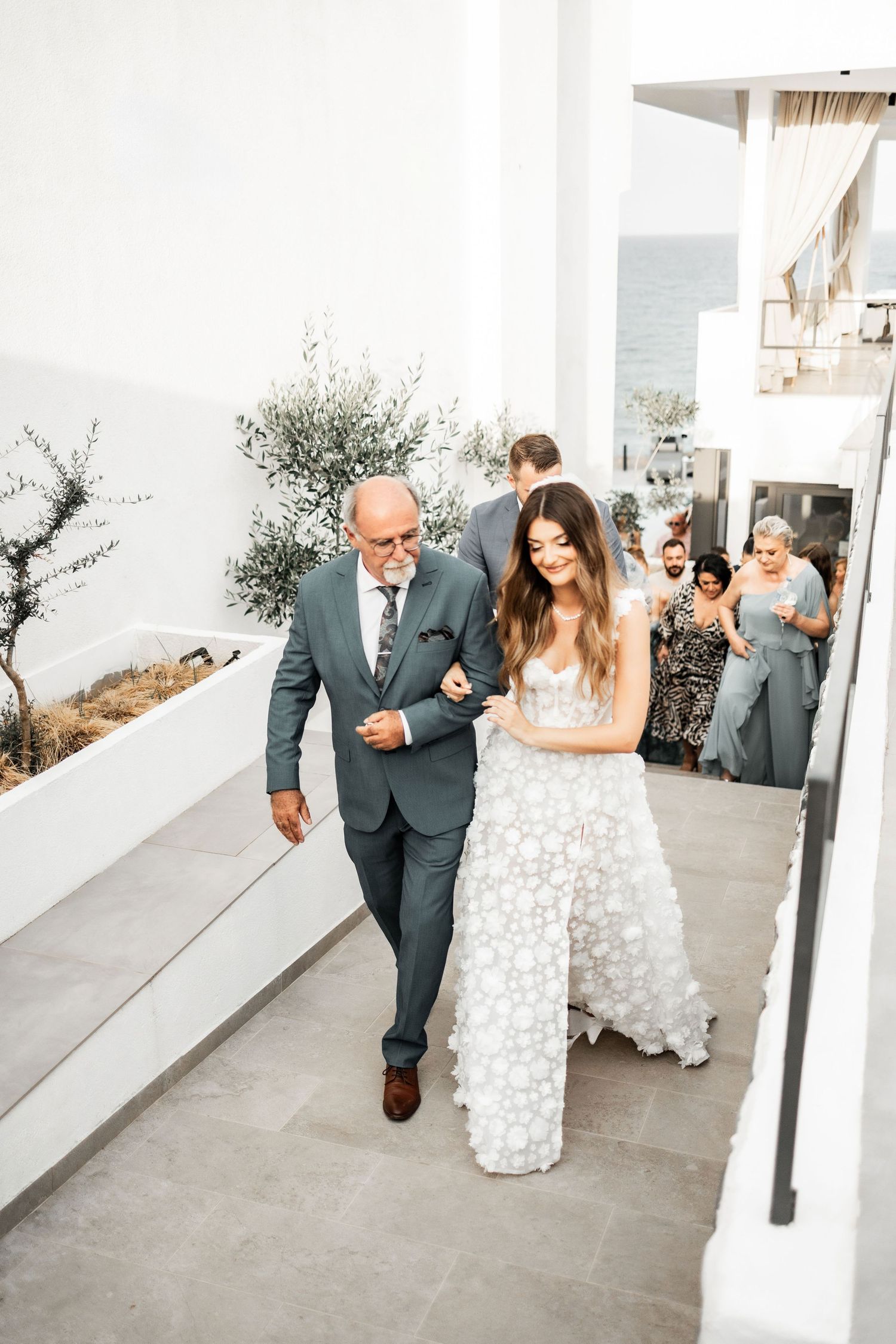 Wedding party walks along a white-walled outdoor pathway.