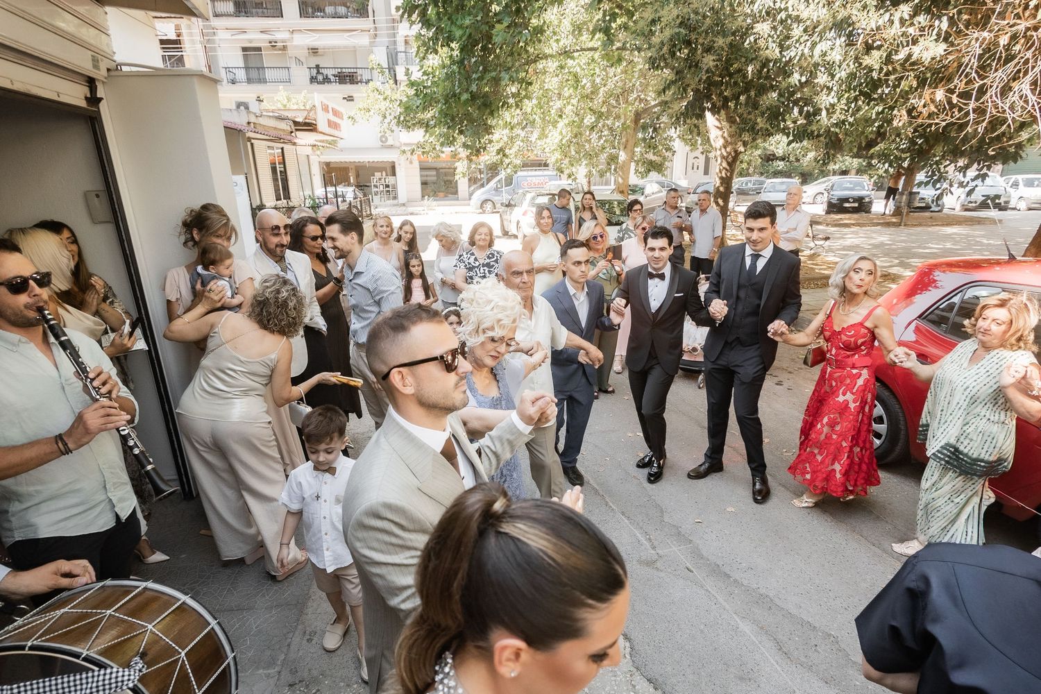 Wedding guests gather outside a venue on a sunny day, with the groom and groomsmen in suits celebrating.