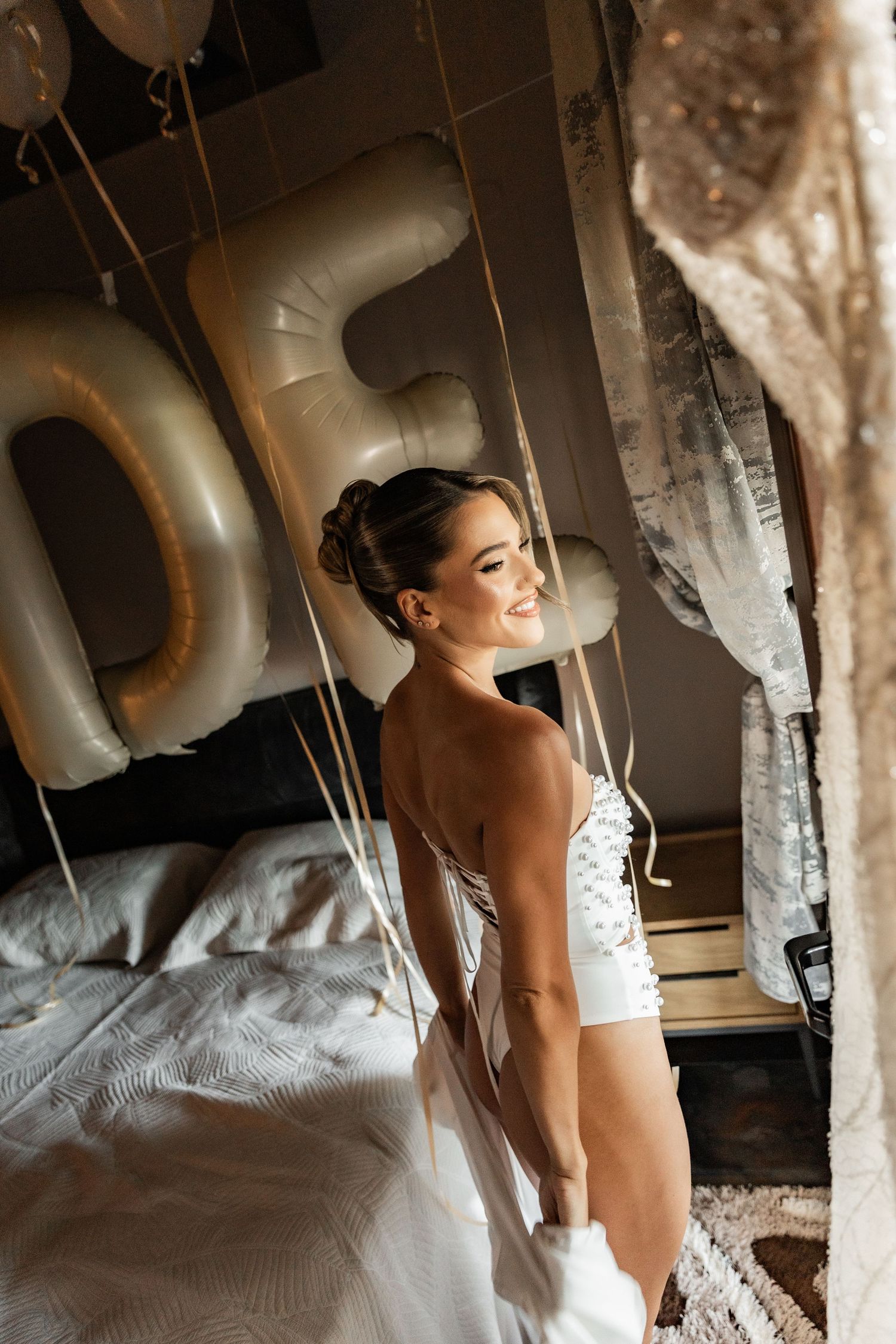Bride in white corset dress smiling in decorated bedroom with gold number balloons on her wedding day.