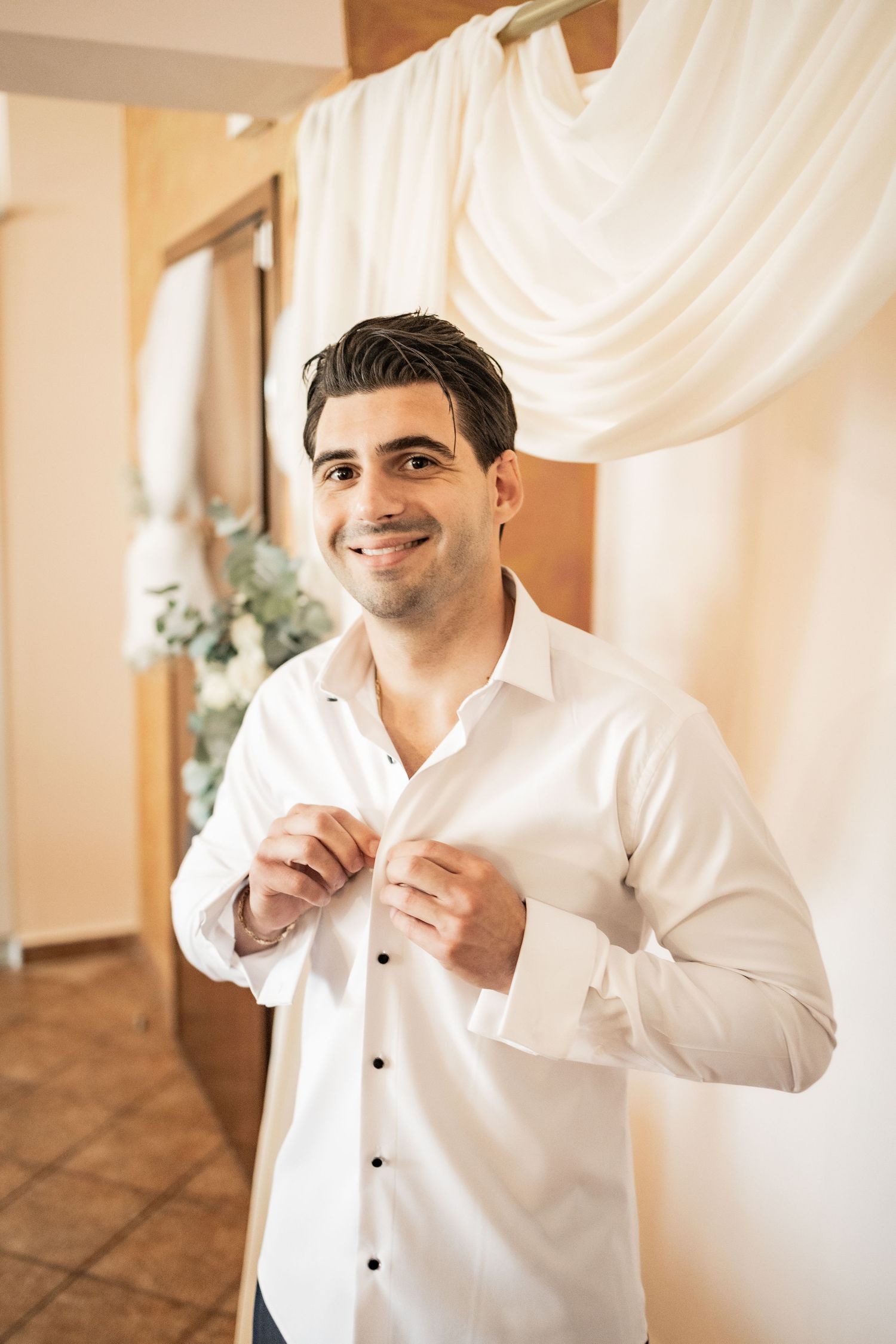 Smiling groom in white dress shirt buttoning up while getting ready for wedding ceremony indoors.