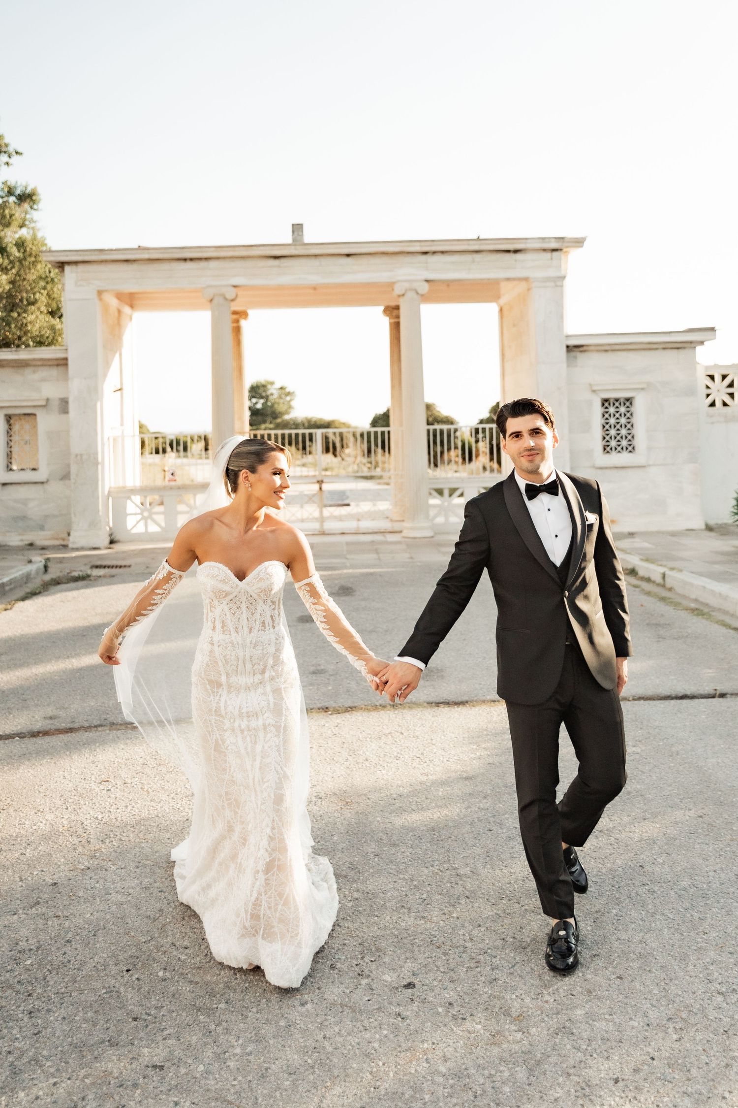 Bride in strapless beaded gown and groom in black tuxedo hold hands walking near classical white columns.