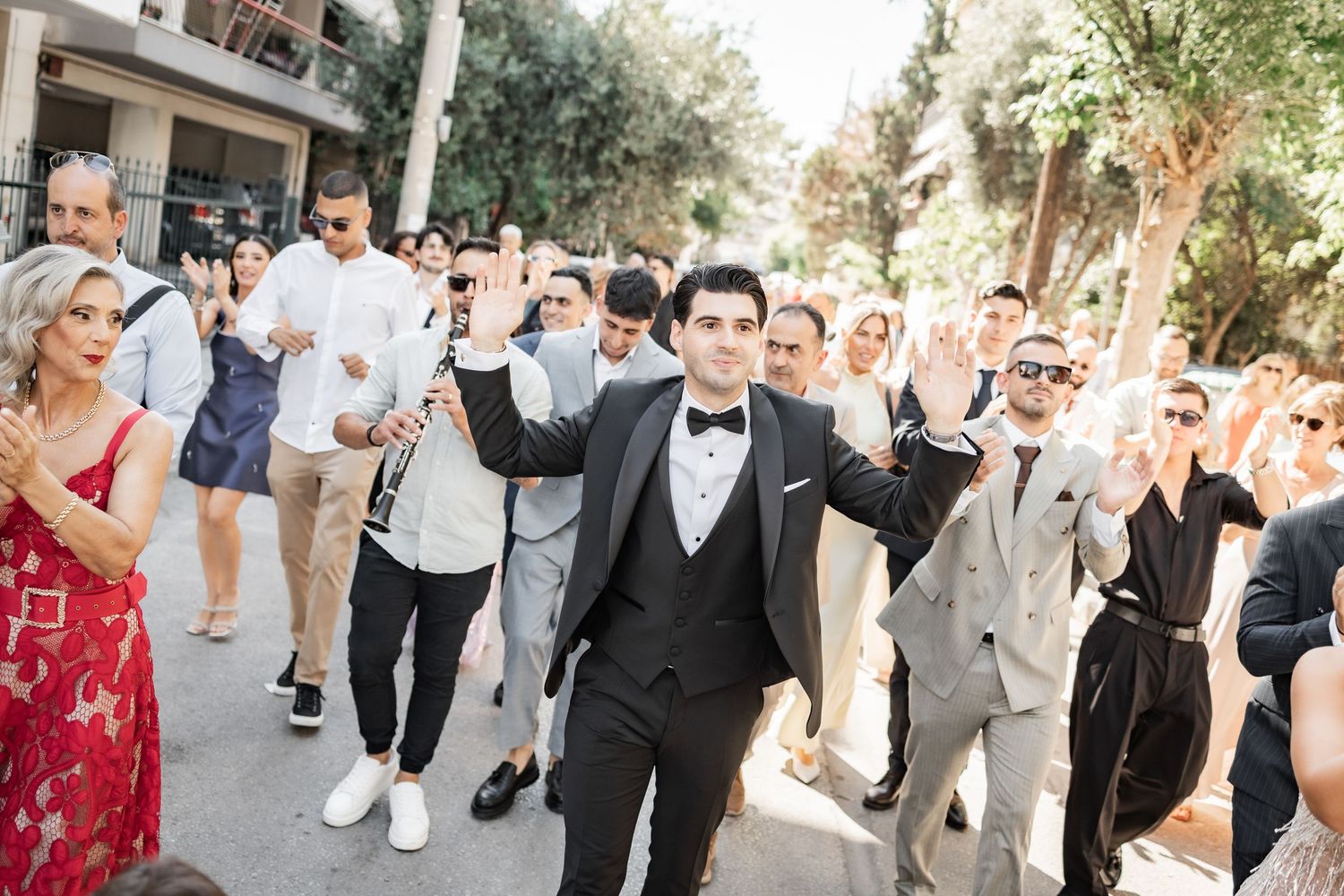 Groom in black tuxedo leads joyful wedding procession outdoors, guests celebrating with raised arms amid lush greenery.