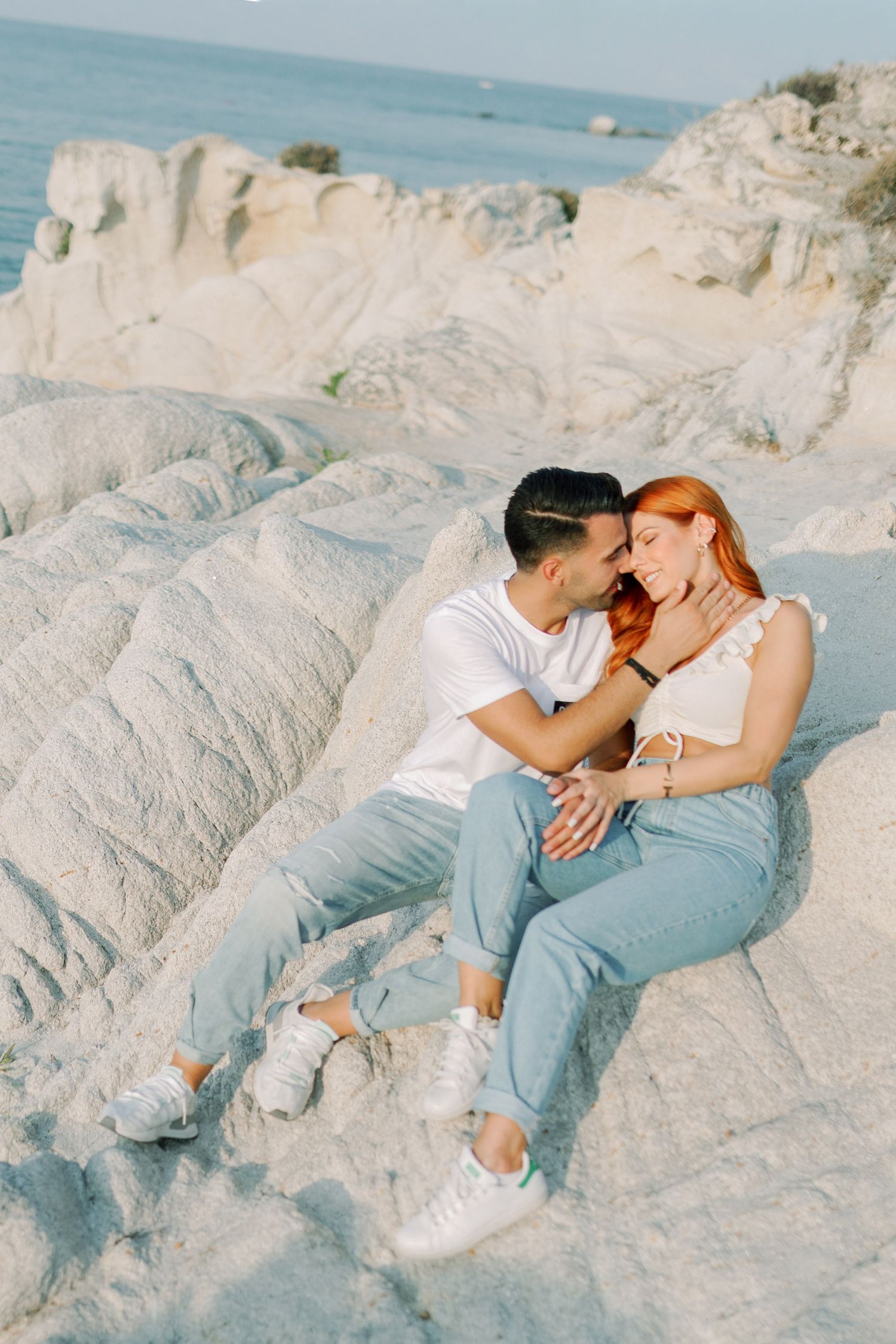 A couple in casual outfits lounging together on white coastal rocks by the ocean on a sunny day.