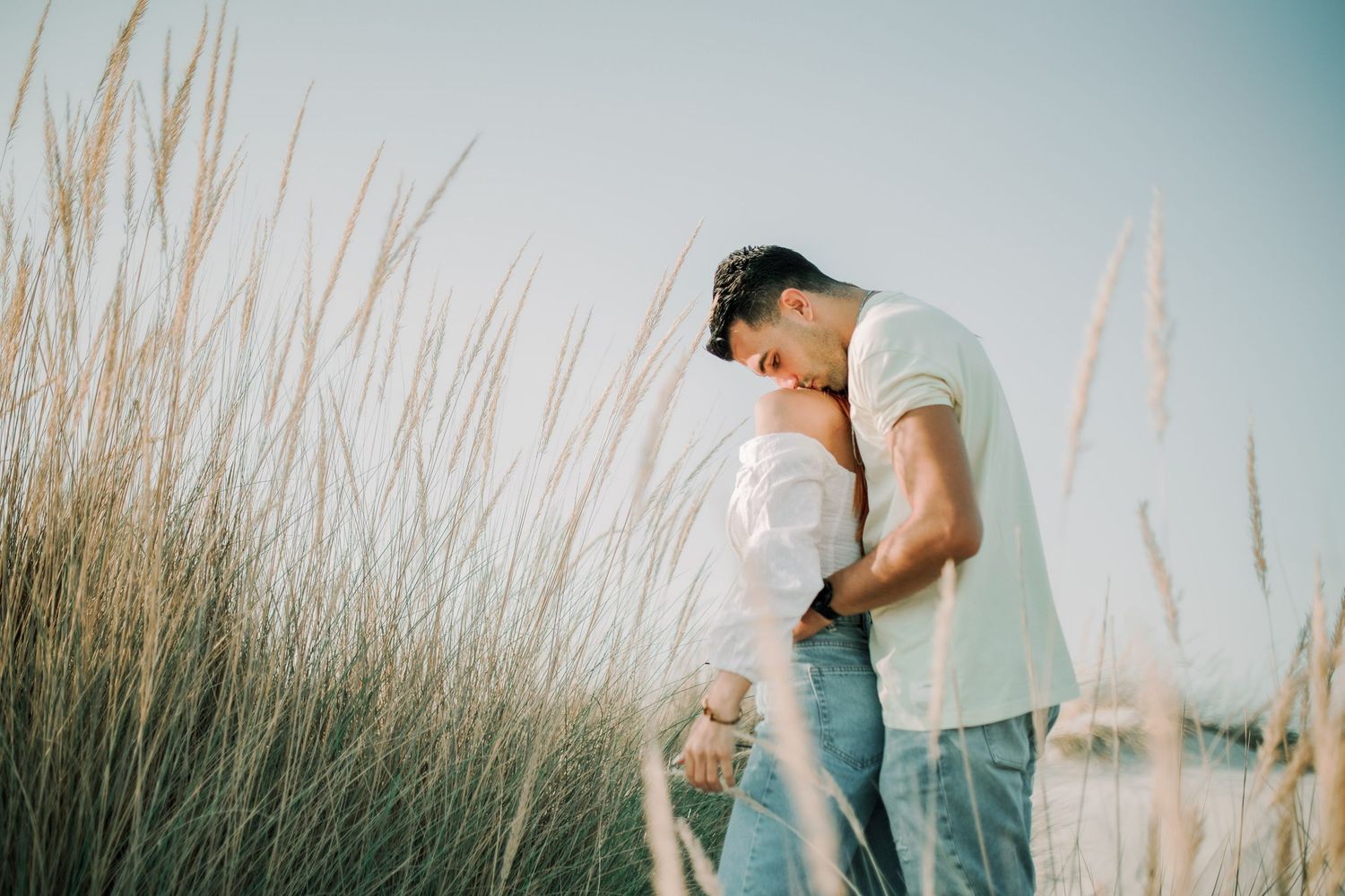 A couple shares a romantic moment in tall grass at sunset, creating a dreamy silhouette against the sky.