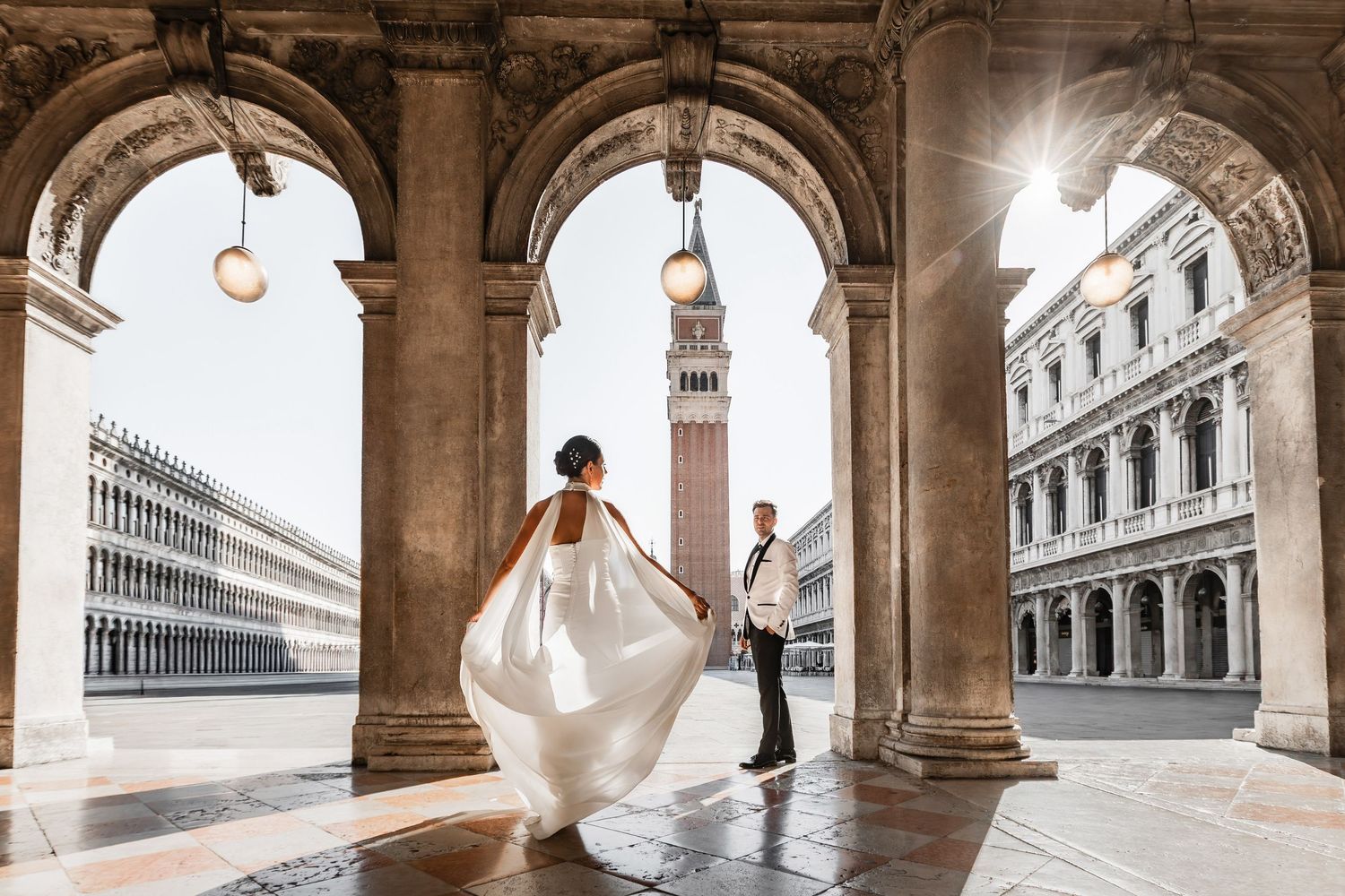 A flowing white dress twirls under historic Venetian arches with the bell tower visible in the distance.