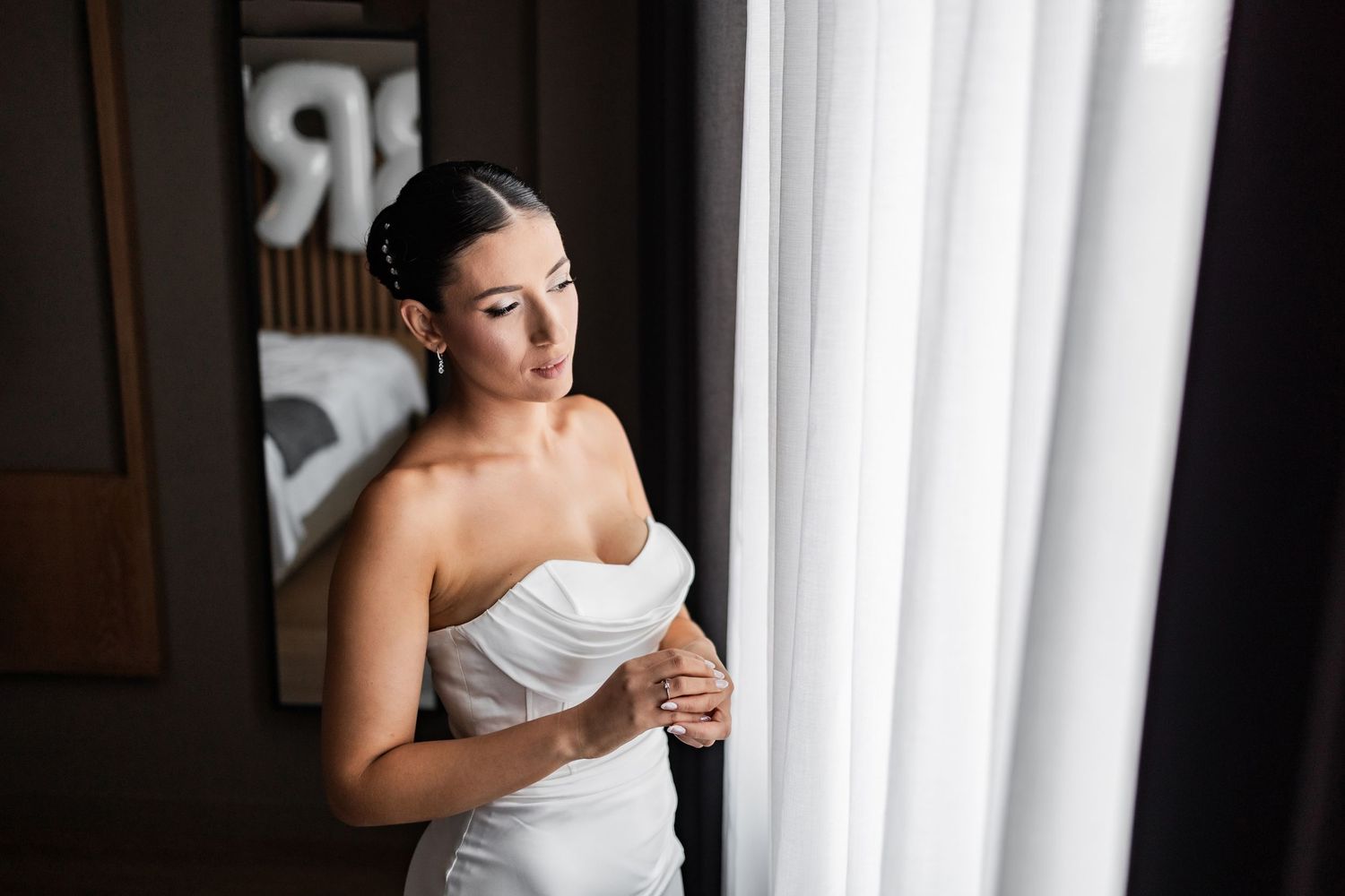 Bride in strapless white gown poses thoughtfully by window with natural light streaming in.