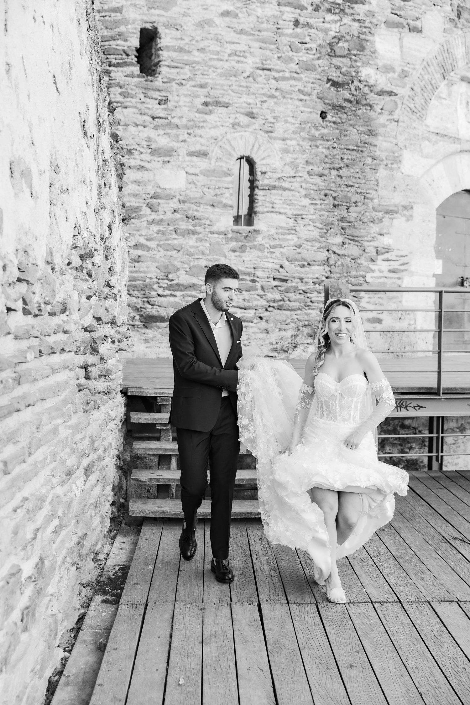 Black and white wedding photo of bride and groom walking on wooden deck beside rustic stone wall.