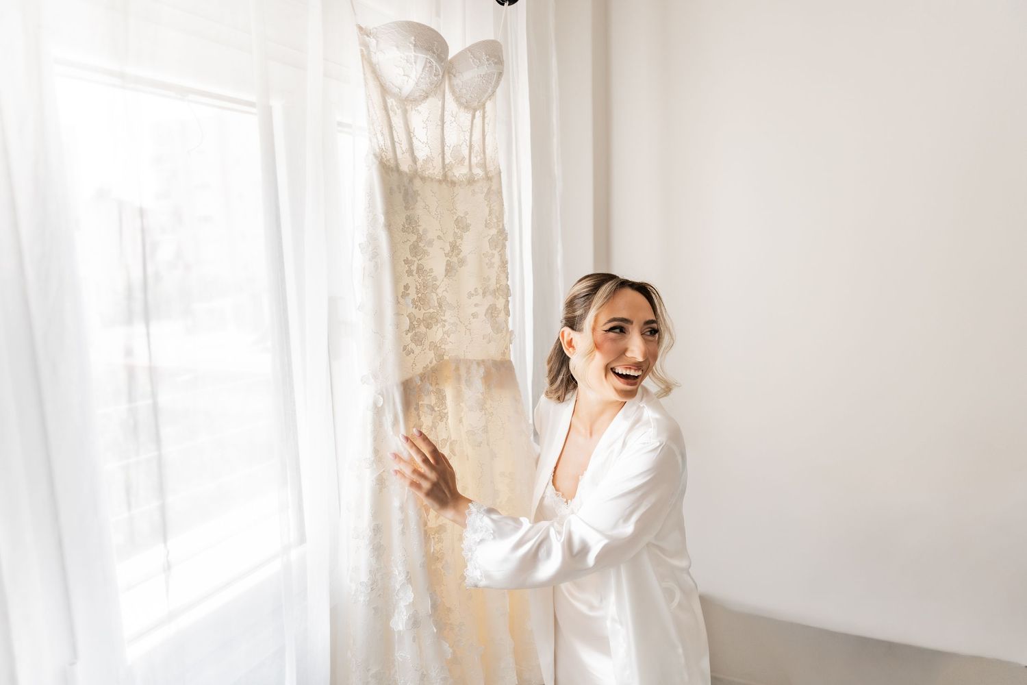 Bride in white robe smiling beside her lace wedding dress hanging by a bright window on her wedding day.
