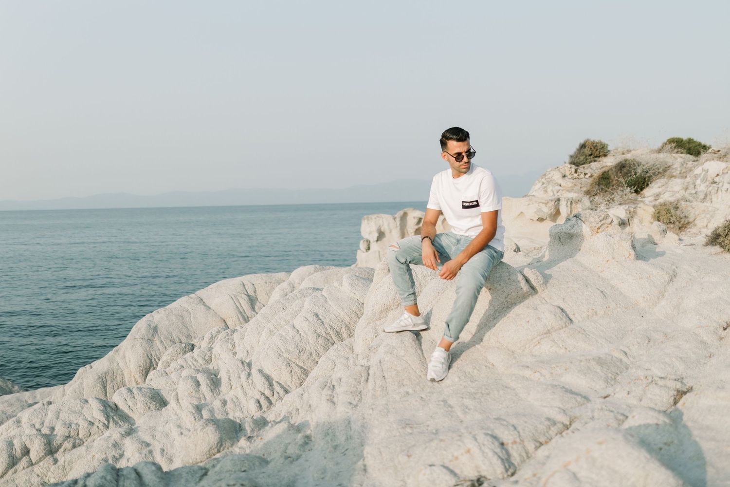 A person in white clothing sits on dramatic white chalk cliffs overlooking the ocean on a sunny day.