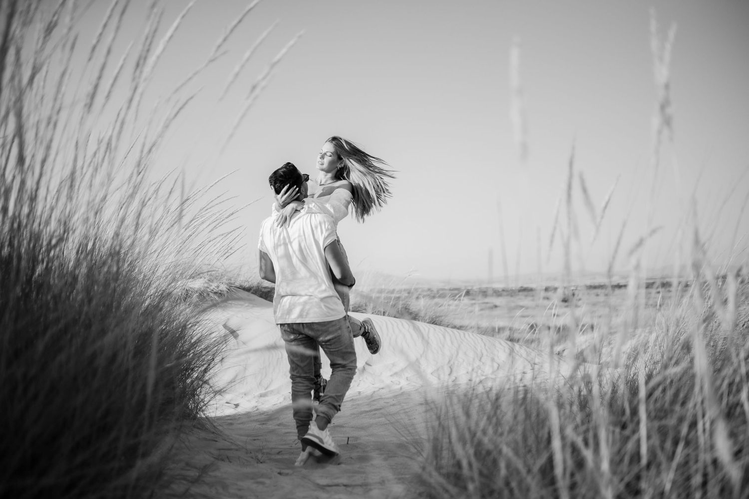 A person runs joyfully through tall beach grass along a sandy path in black and white.