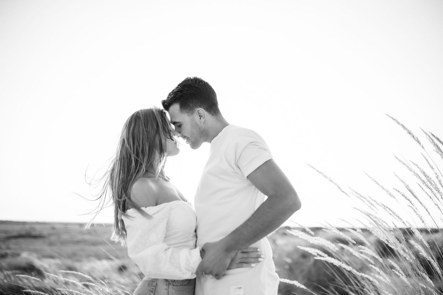 A romantic black and white photo of a couple embracing in tall grass against a bright sky.