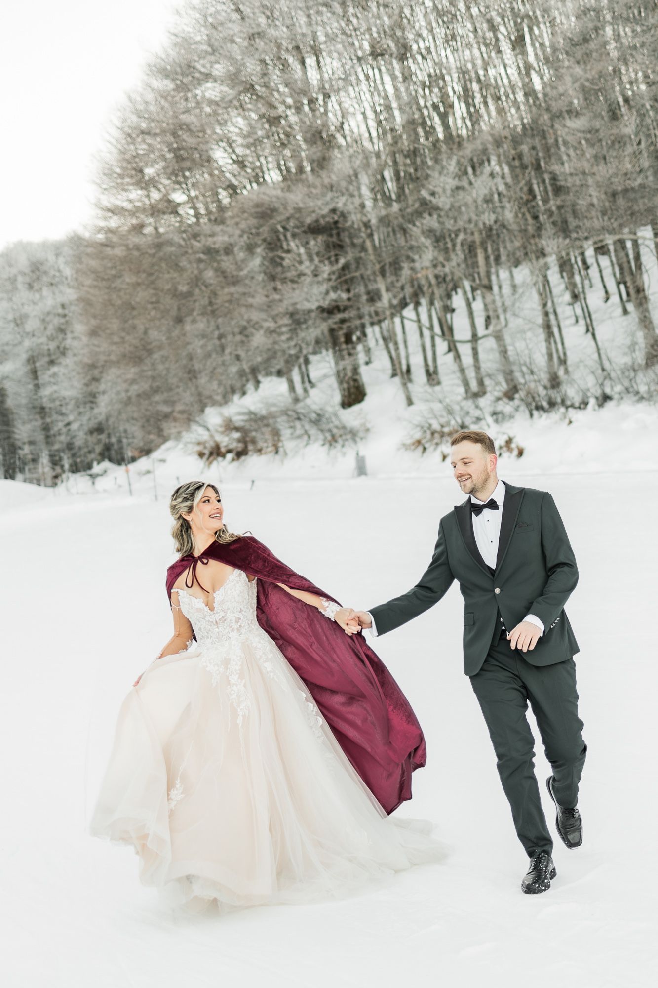 A couple plays in the snow during their winter wedding photoshoot wearing burgundy and formal attire.