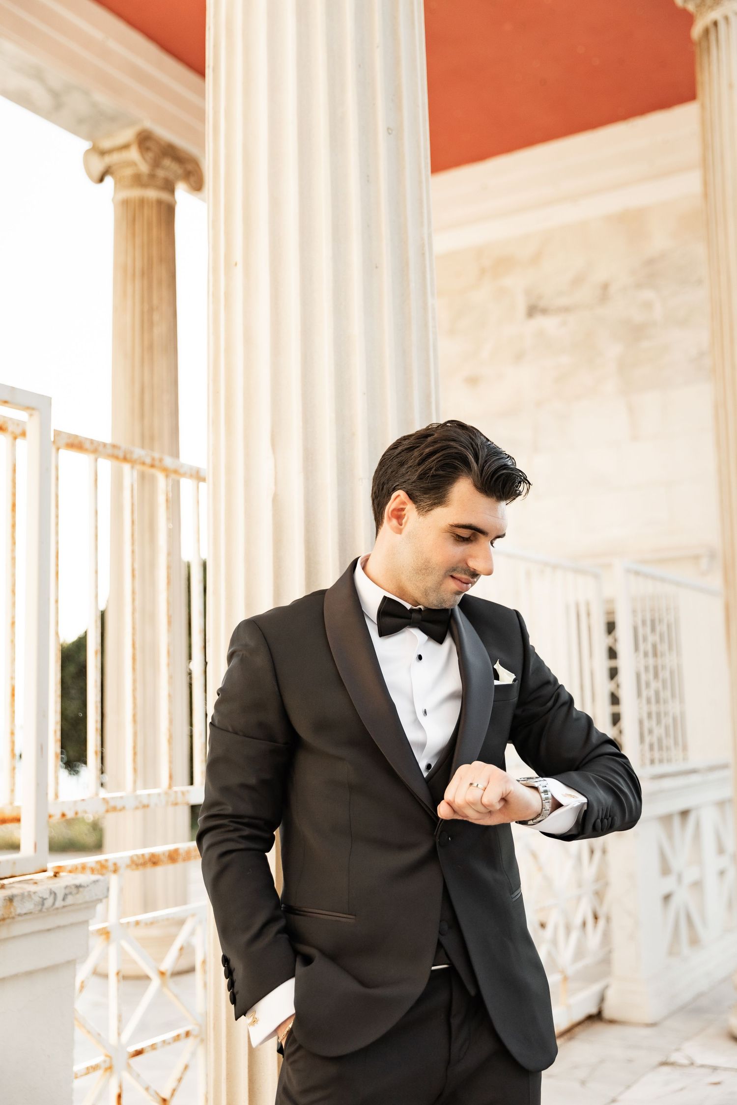 Elegant groom in black tuxedo and bow tie checking his watch beside classical white columns.