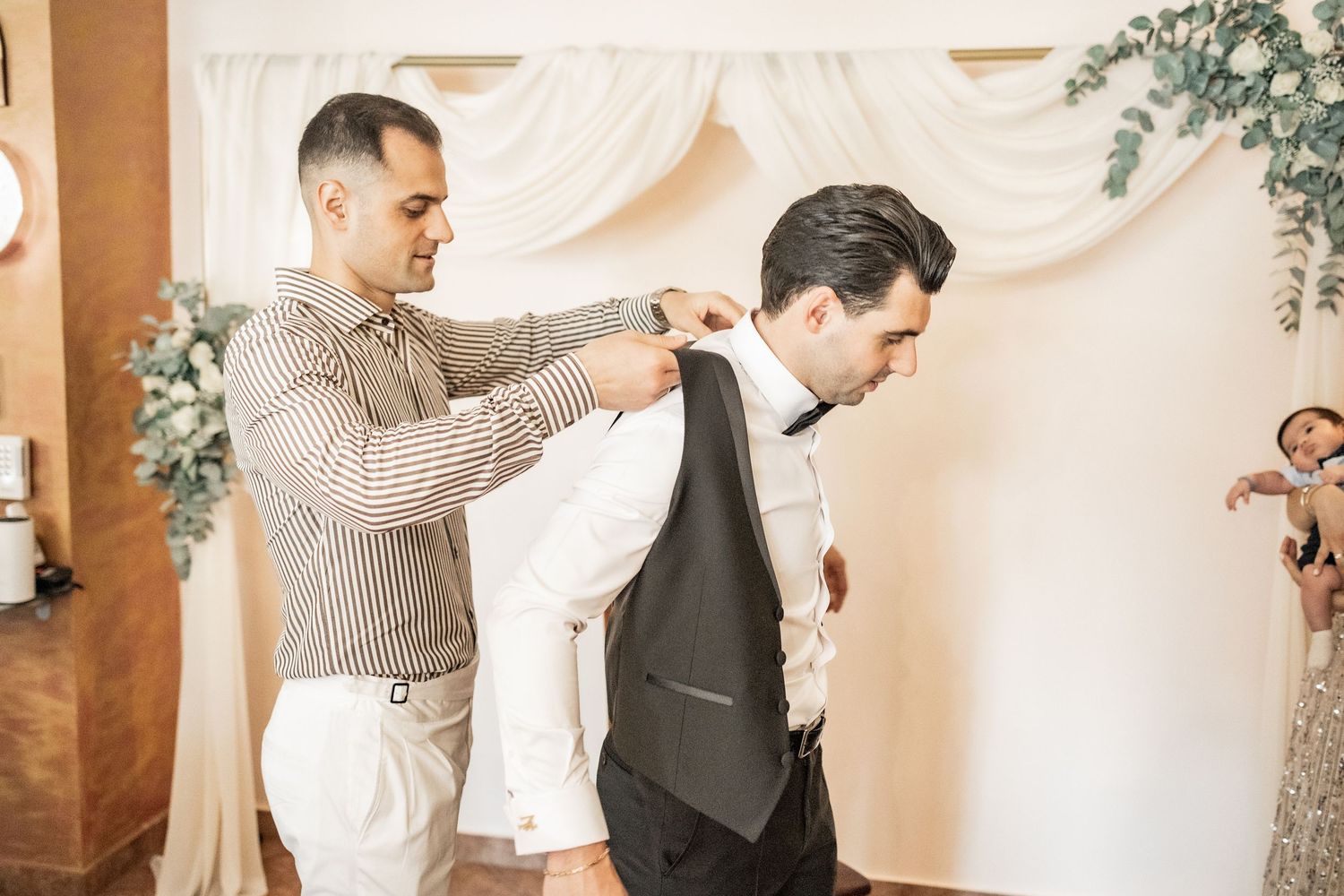 A man helps another man put on a dark suit jacket in an elegant wedding venue with floral decorations.