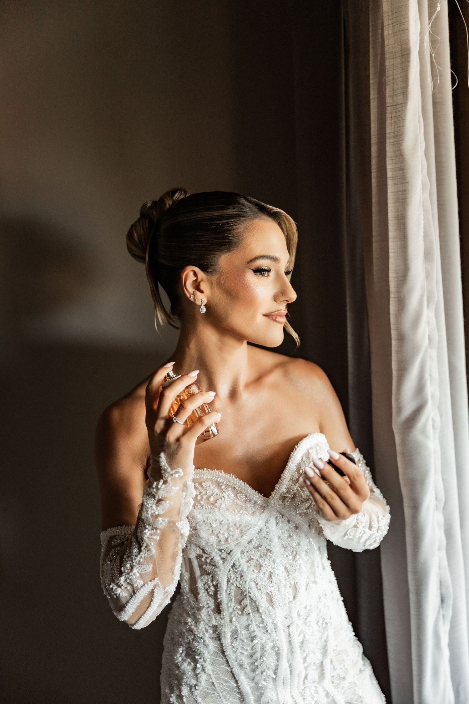 Elegant bride in white lace off-shoulder gown adjusting dress near window, wearing gold rings and pearl earrings.