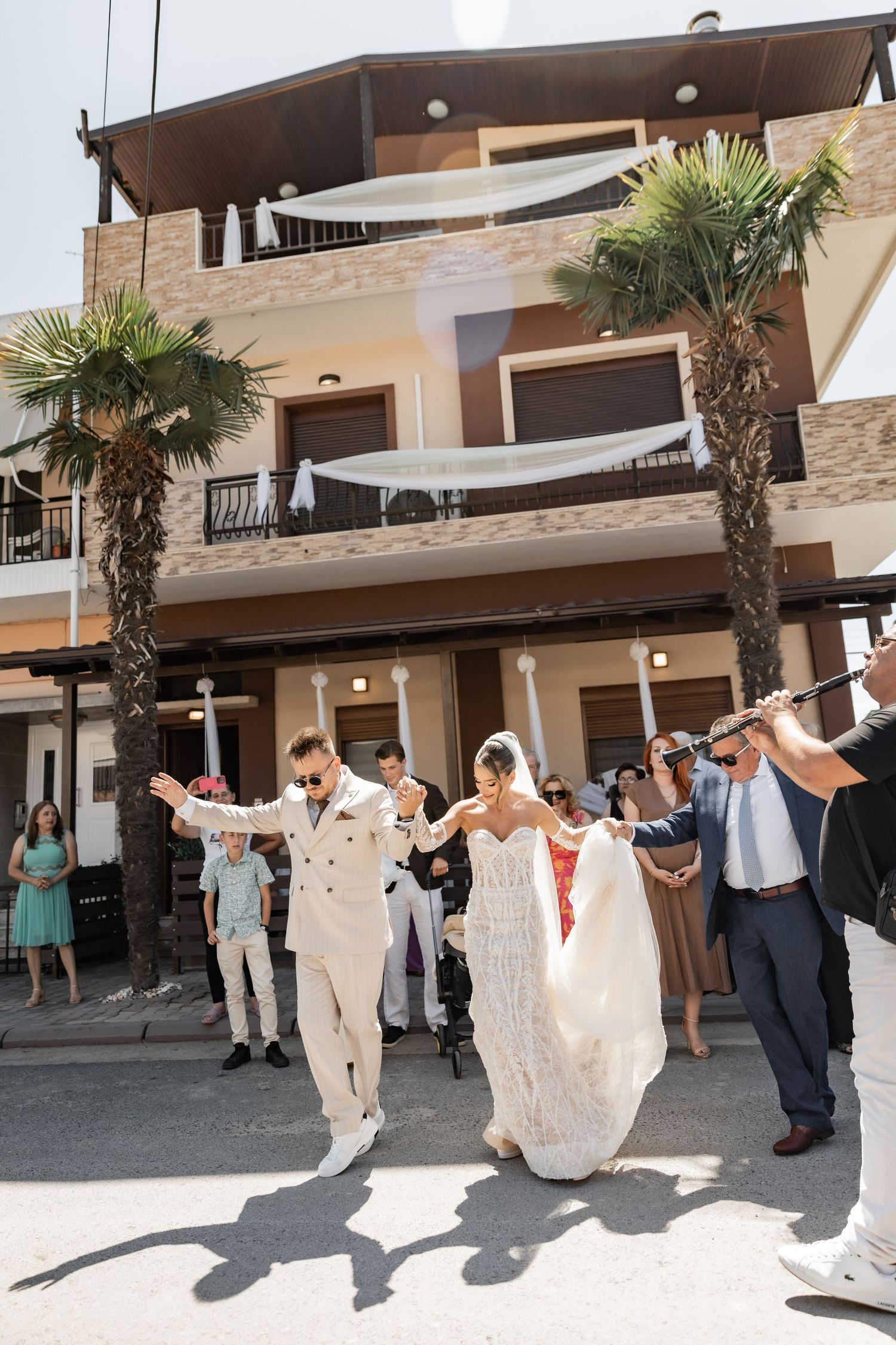 Bride and groom dancing joyfully outside a Mediterranean building with palm trees, surrounded by wedding guests.