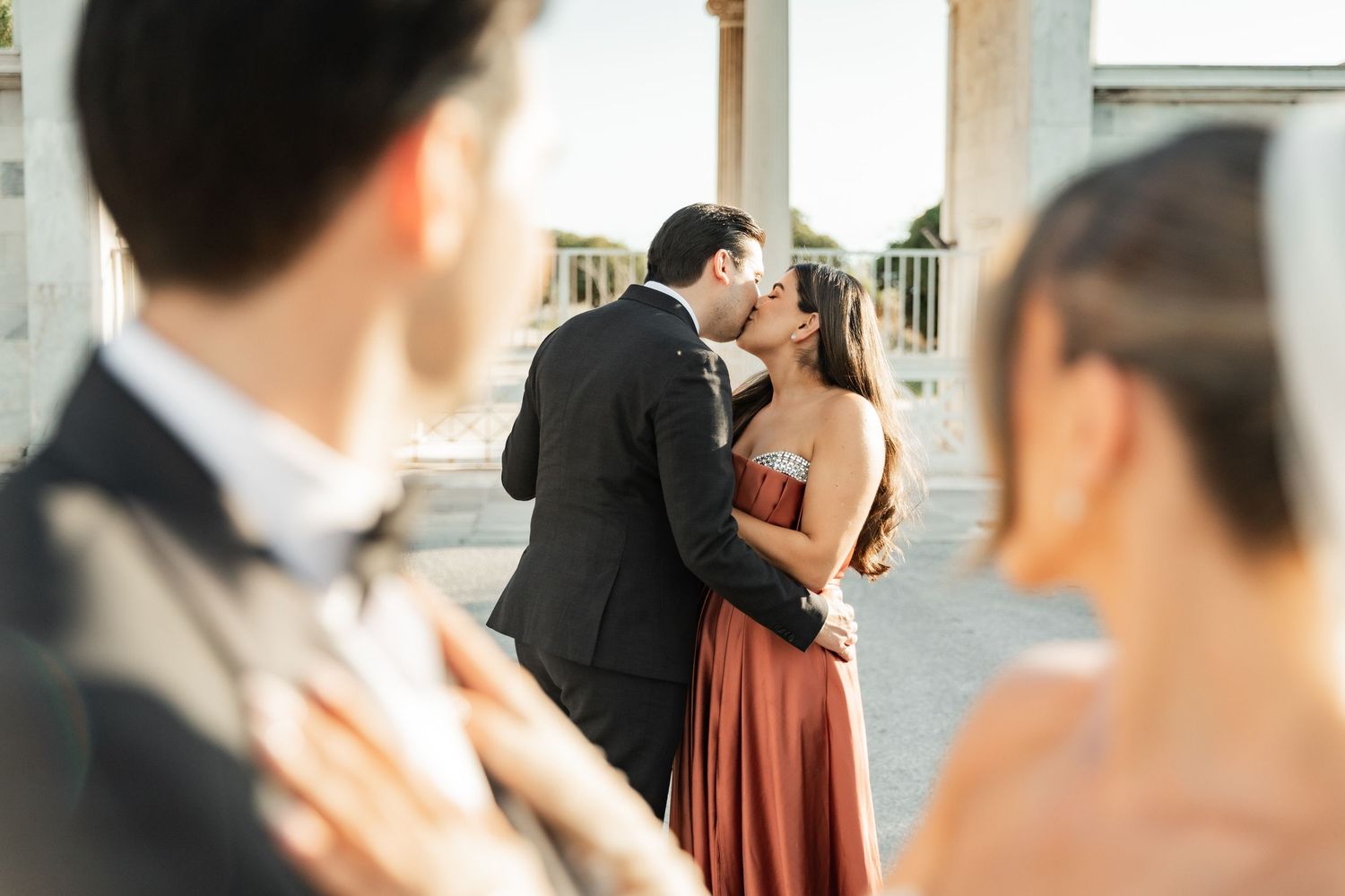 Couple sharing a romantic kiss at an outdoor wedding venue, bride in rust satin gown, groom in dark suit.