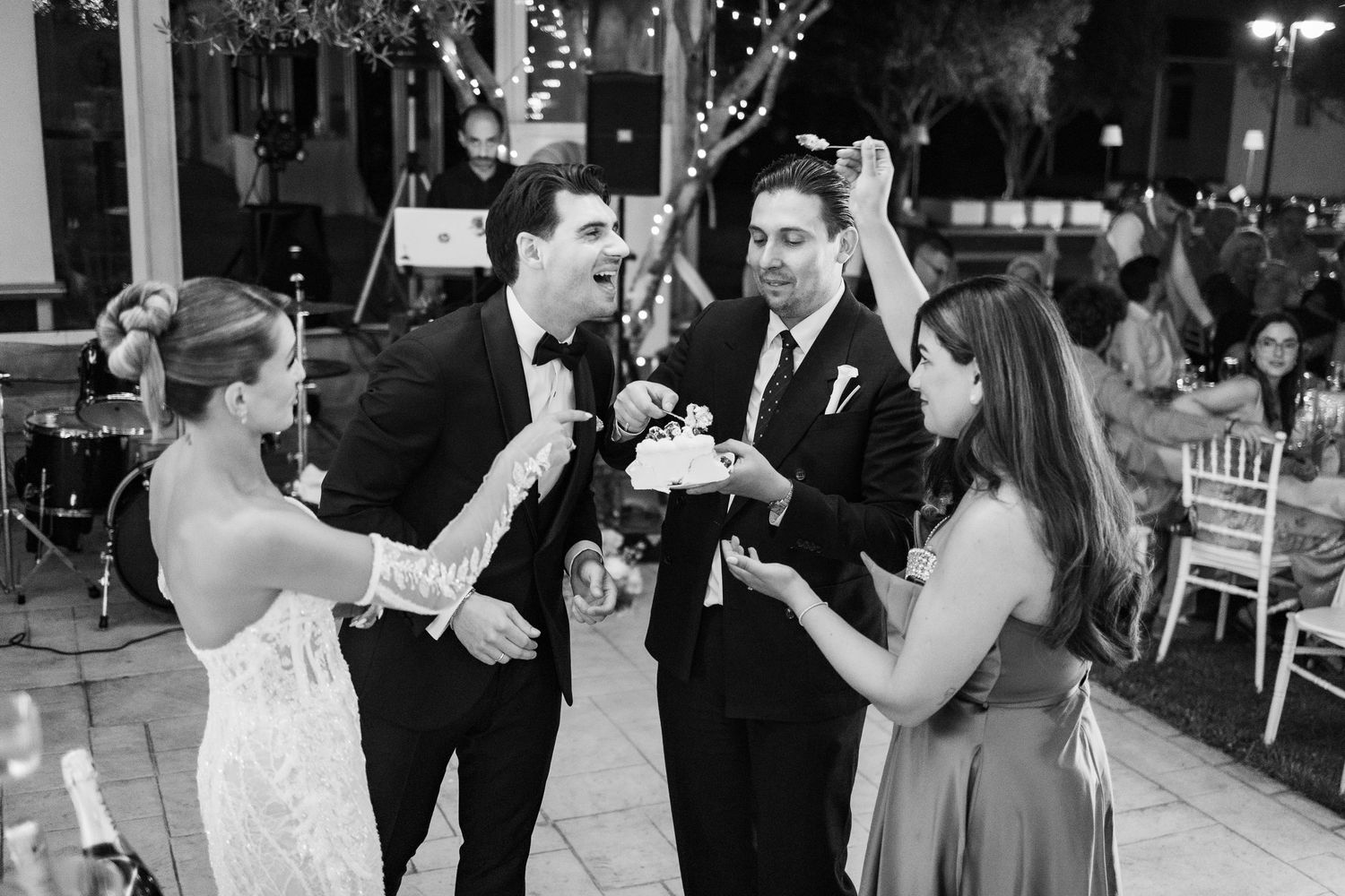 Black and white wedding reception photo of couple cutting cake with guests celebrating on outdoor patio.